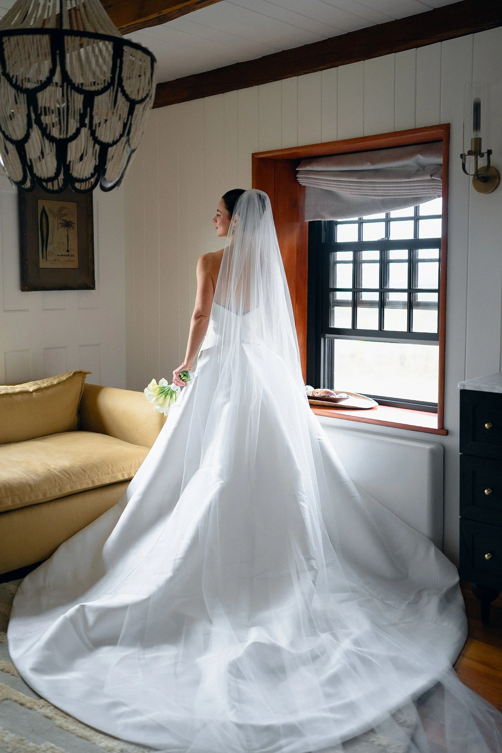 Bride in a white wedding gown with a long train and veil, holding a bouquet, standing indoors near a window with wooden trim.