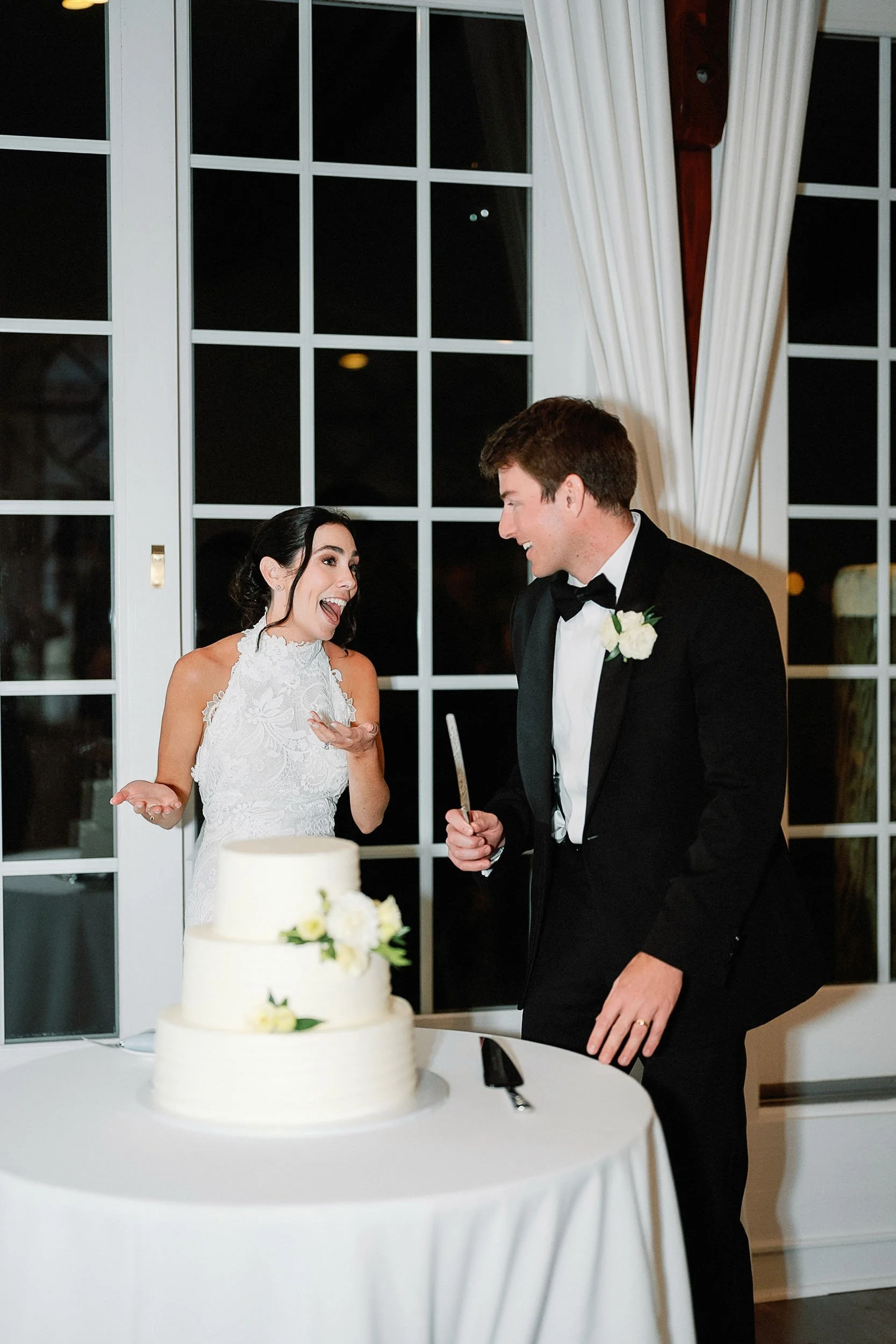 A bride and groom celebrating their wedding cake, smiling and laughing indoors at night.