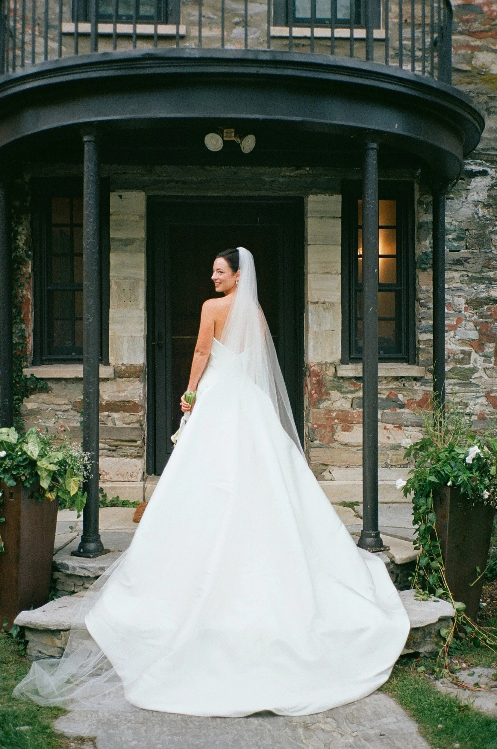 A bride in a white wedding gown with a long train and veil, standing in front of a rustic stone and brick house with black windows and a black door, holding a small bouquet, looking over her shoulder and smiling.