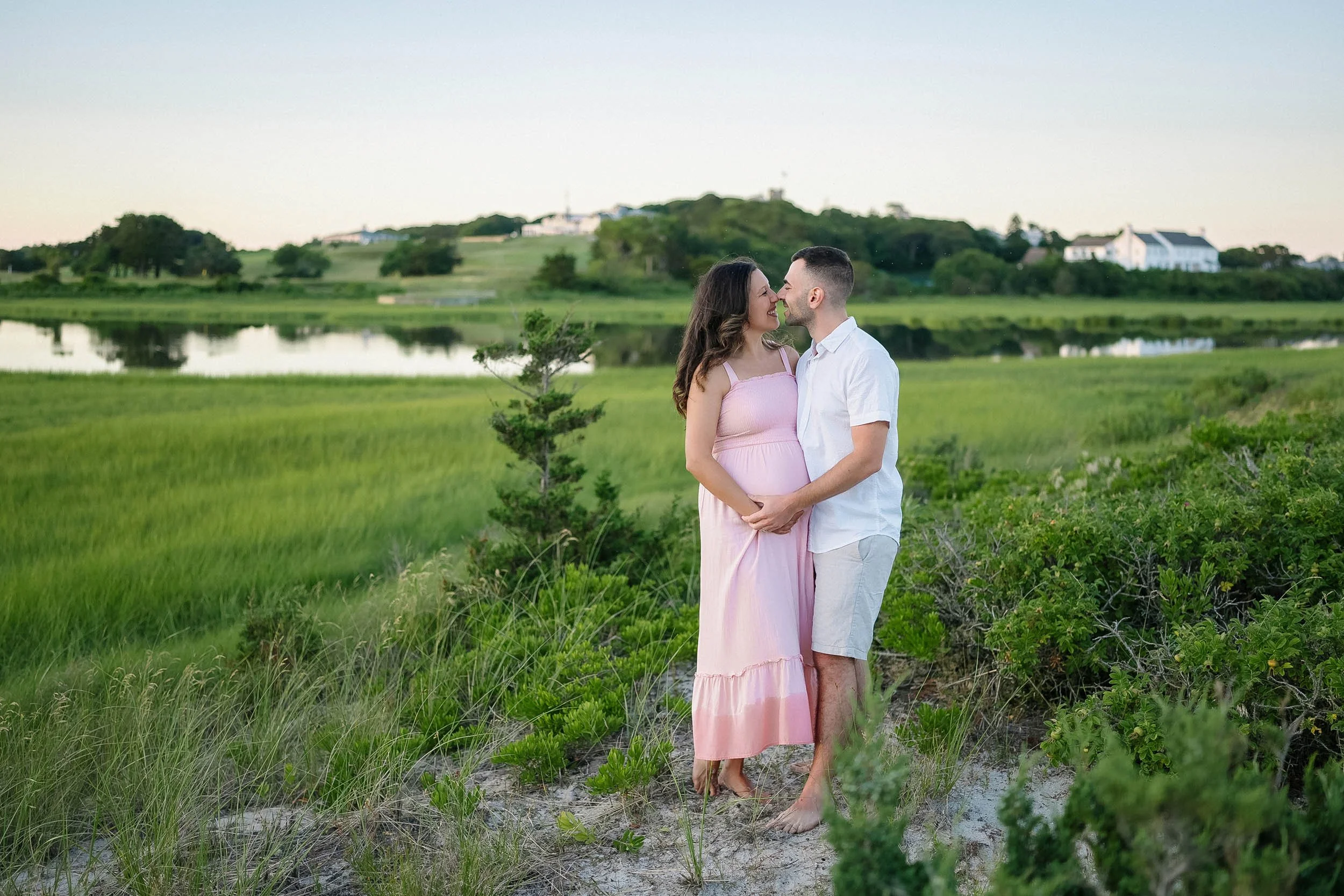 A couple stands close together on a grassy path near a lake and lush green landscape at sunset, smiling and about to kiss.