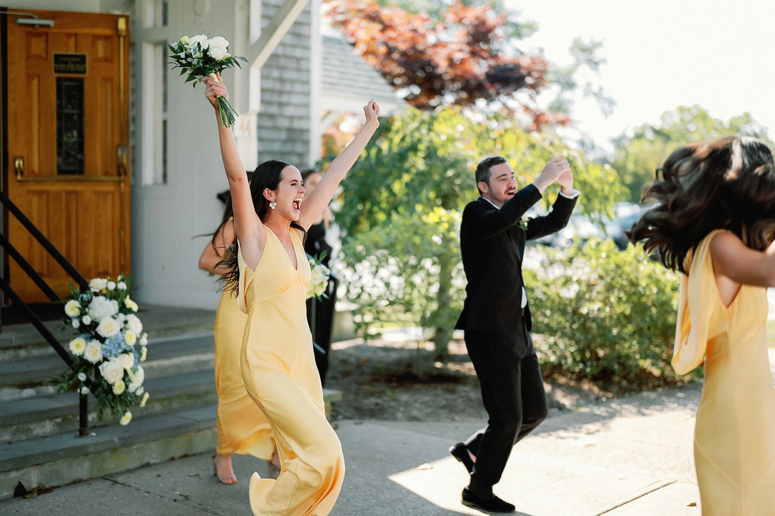 Joyful wedding celebration outside, woman in yellow dress holding a bouquet with both arms raised, man in black suit dancing, sunny day with trees and a decorated building entrance in background.