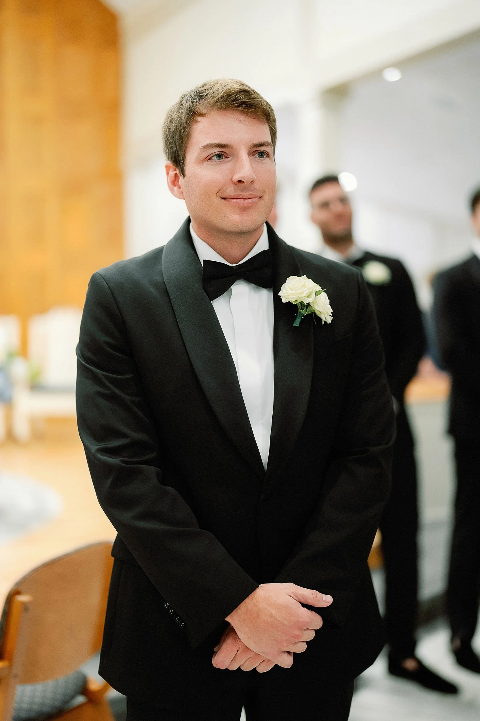 A groom in a black tuxedo with a boutonnière, standing with hands clasped, at a wedding ceremony.