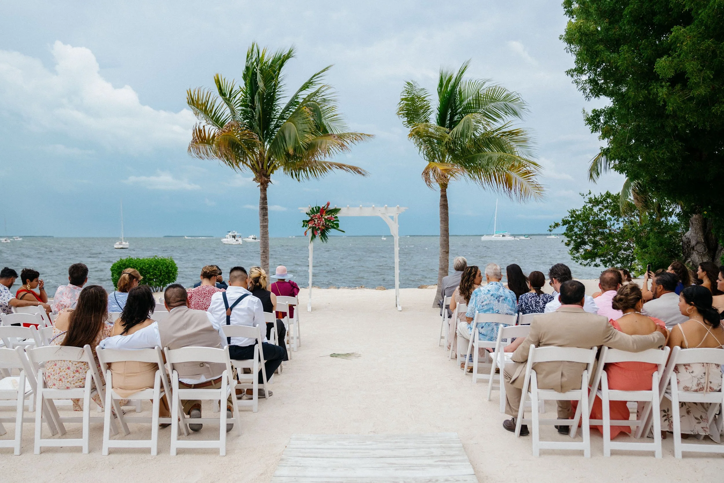 Wedding ceremony setup on a beach with chairs arranged in two rows, a white arbor decorated with flowers, palm trees, and boats on the water in the background.
