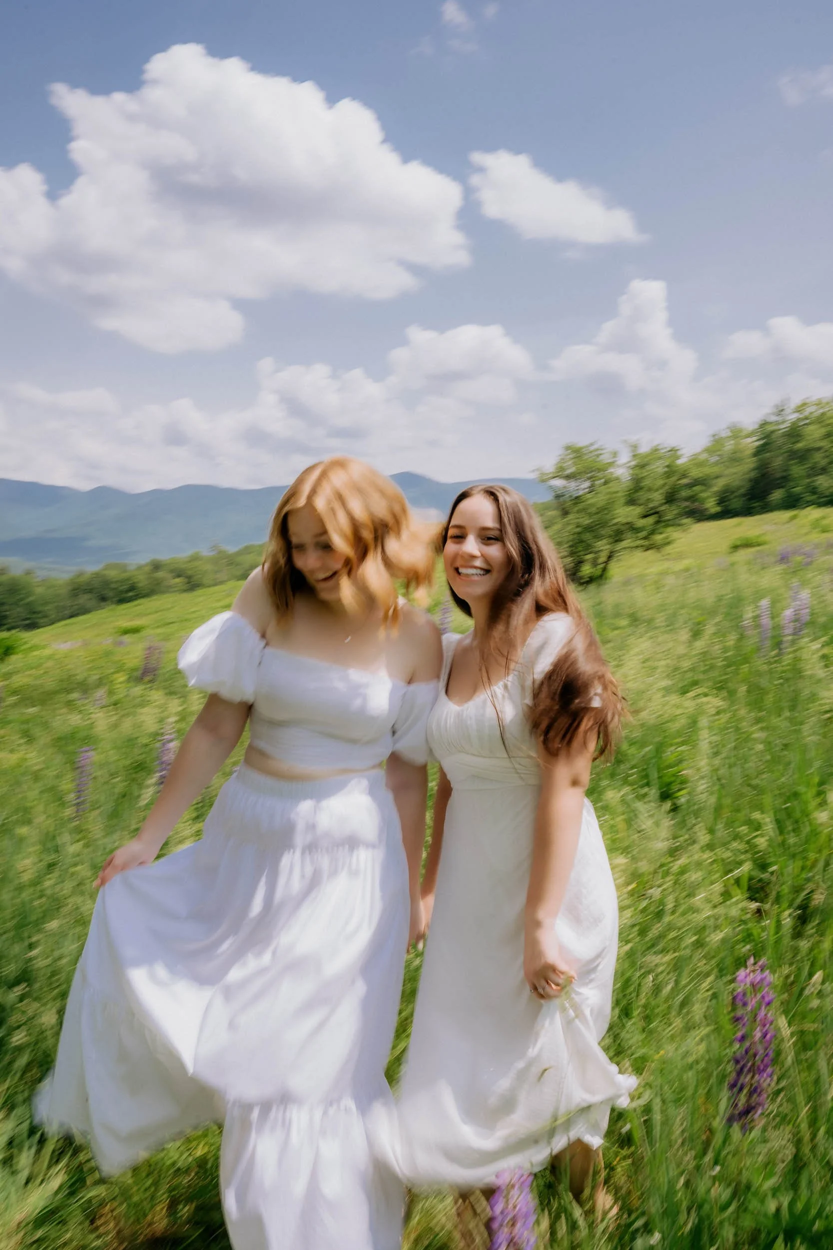 Two smiling women in white dresses walk through a green field with purple flowers, under a partly cloudy blue sky.