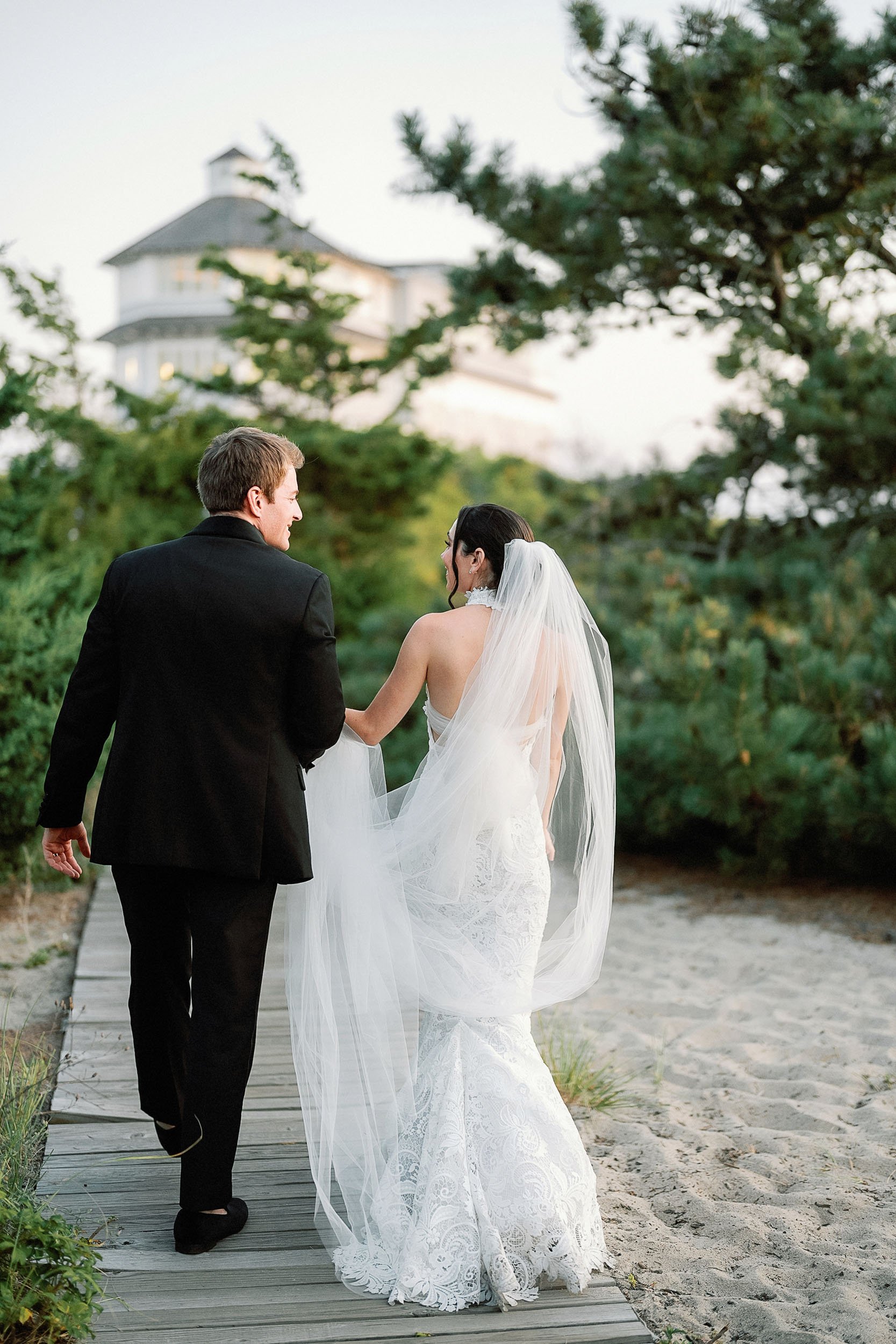 A bride and groom walking on a wooden pathway outdoors, with the bride wearing a white lace wedding dress and veil, and the groom in a black suit, surrounded by greenery and trees, with a large house visible in the background.