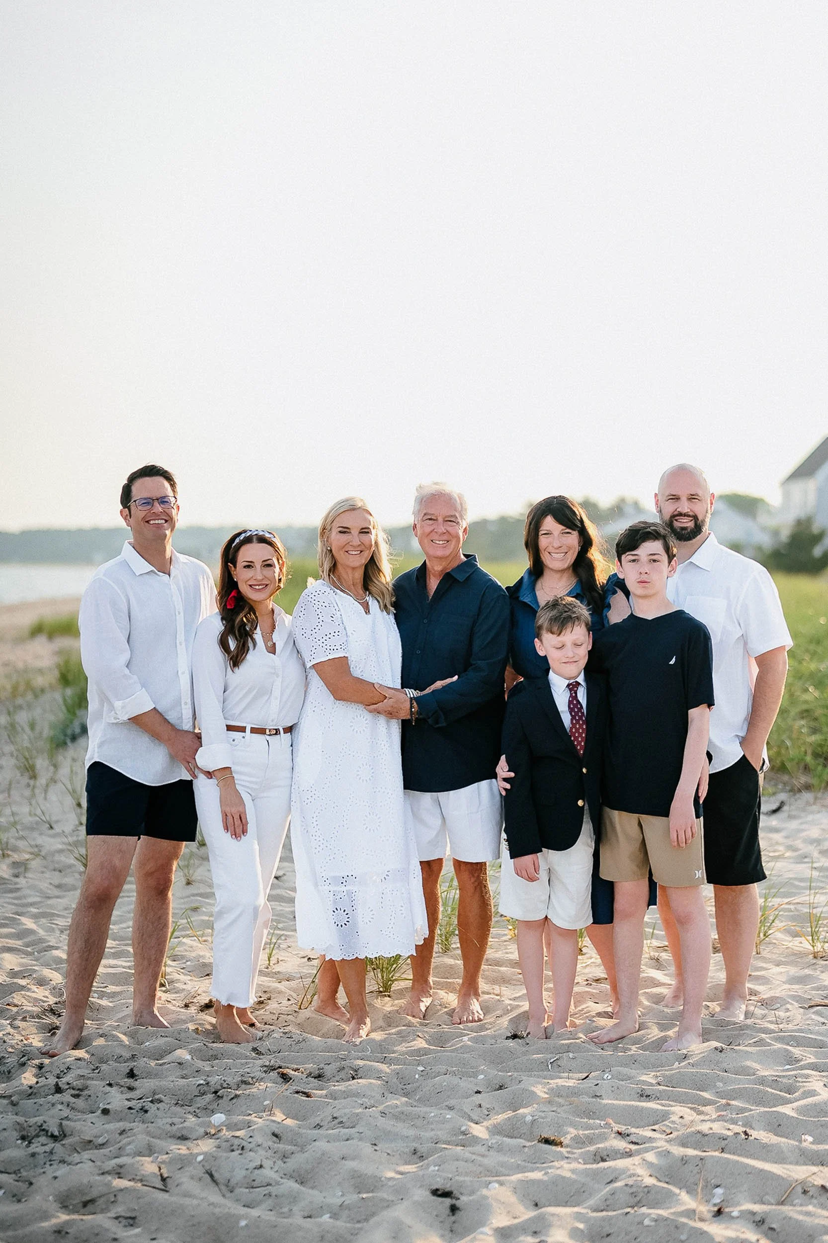 A family of nine standing on a sandy beach, dressed in casual summer clothing, smiling at the camera, with grassy dunes and a house in the background during sunset.