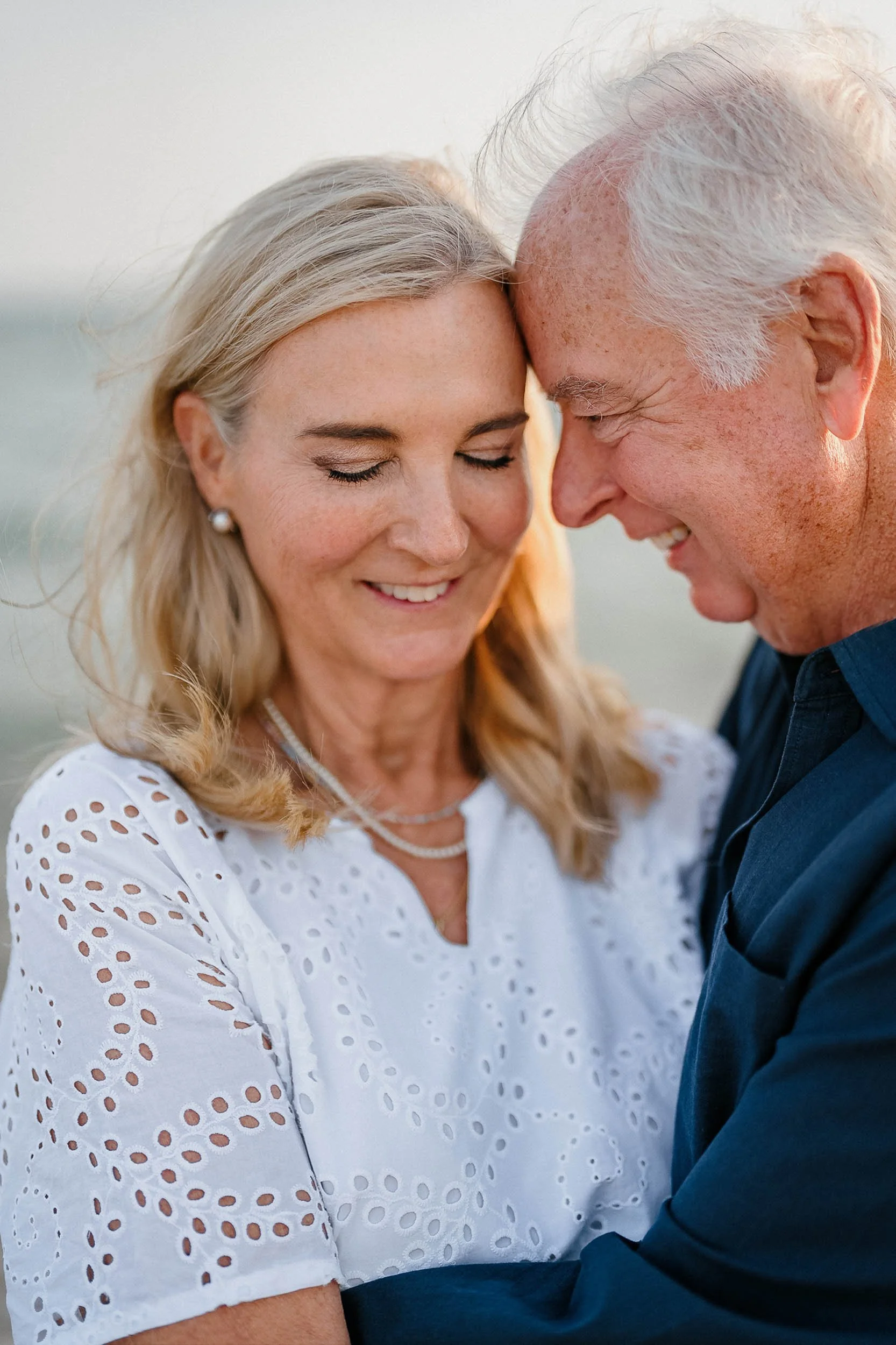 A close-up of a joyful elderly couple with their foreheads and noses touching, eyes closed, smiling, and embracing outdoors during sunset.