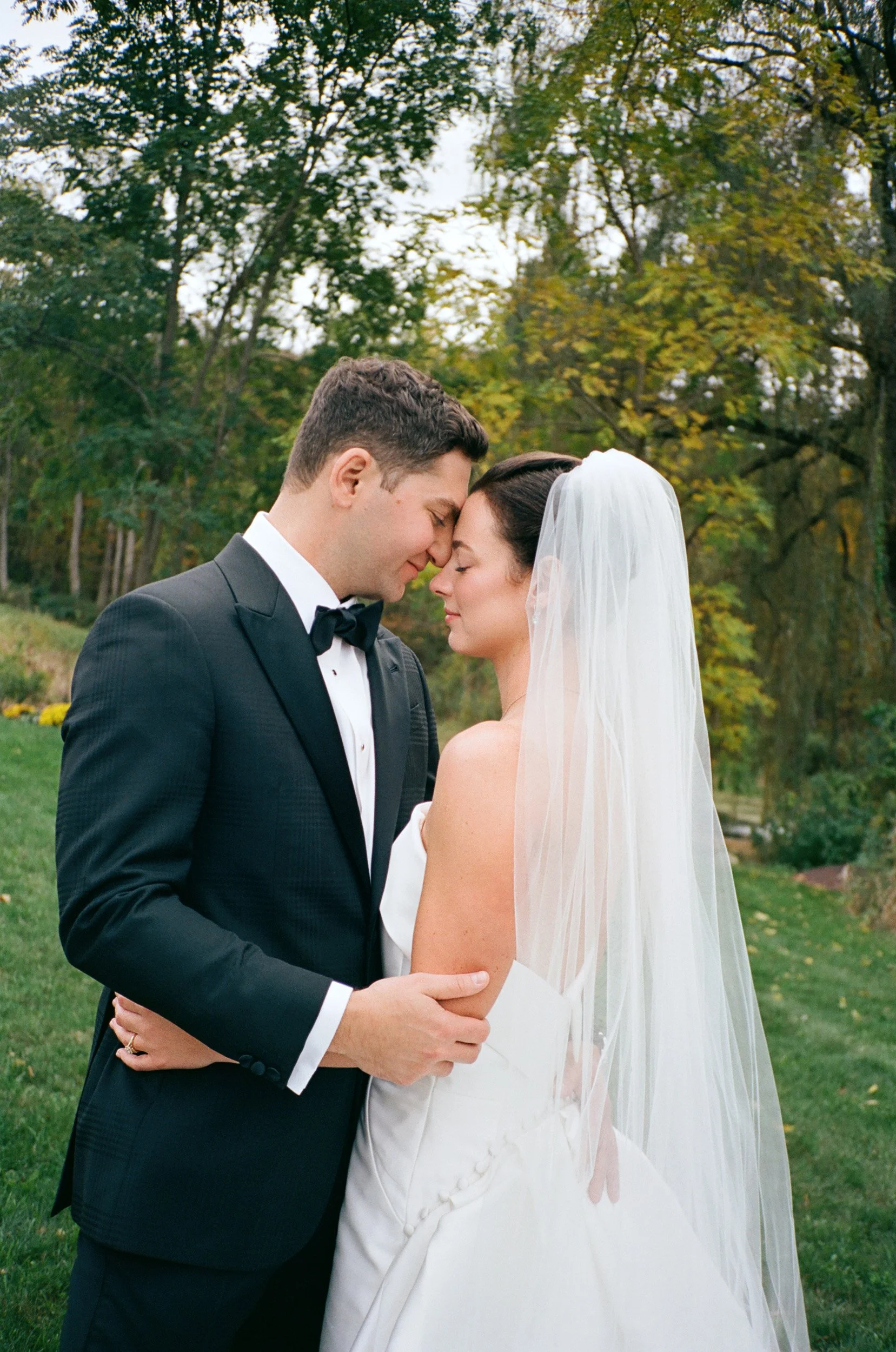 A bride and groom stand close, touching foreheads with their eyes closed, outdoors on a grassy area with trees in the background, during their wedding.