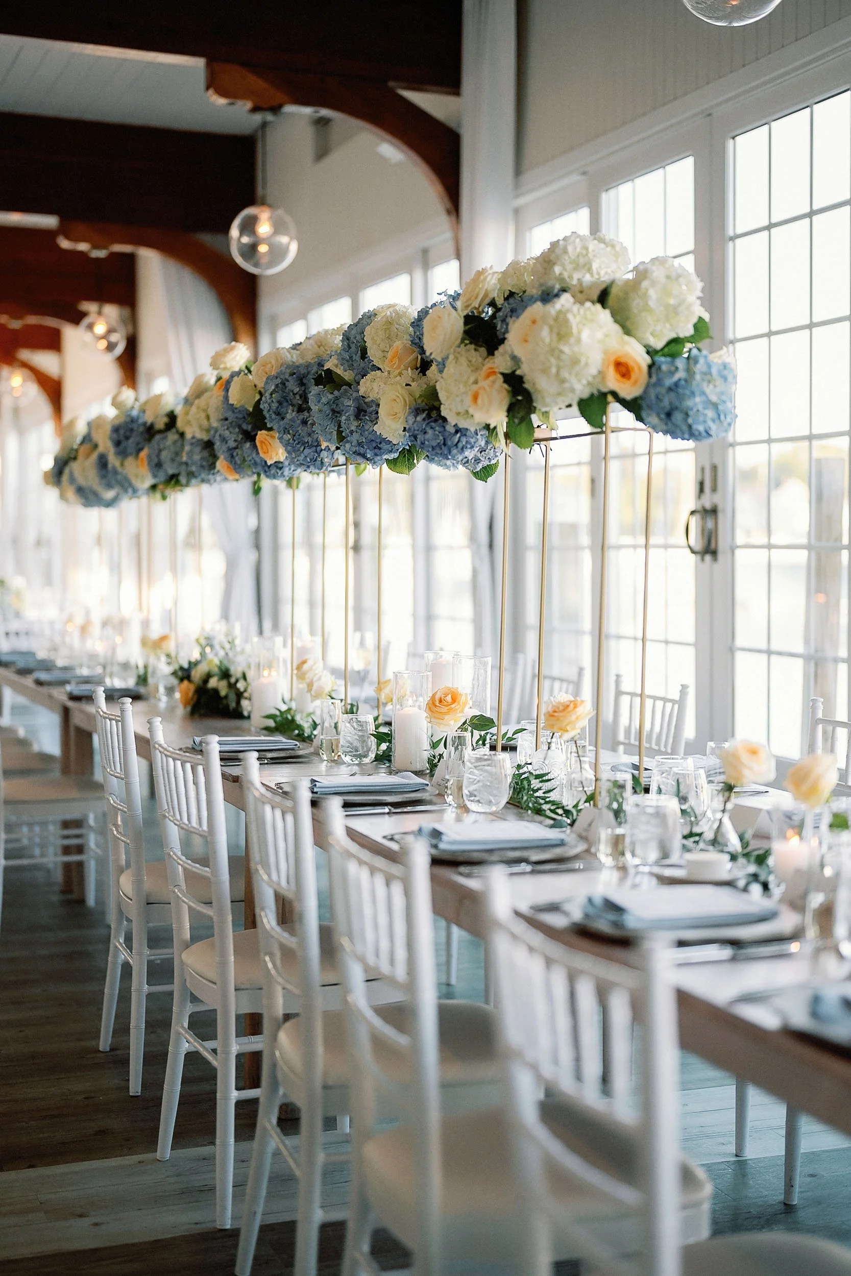 Elegant indoor wedding reception table with a centerpiece of white and blue flowers, candles, and tableware, illuminated by large windows and pendant lights.