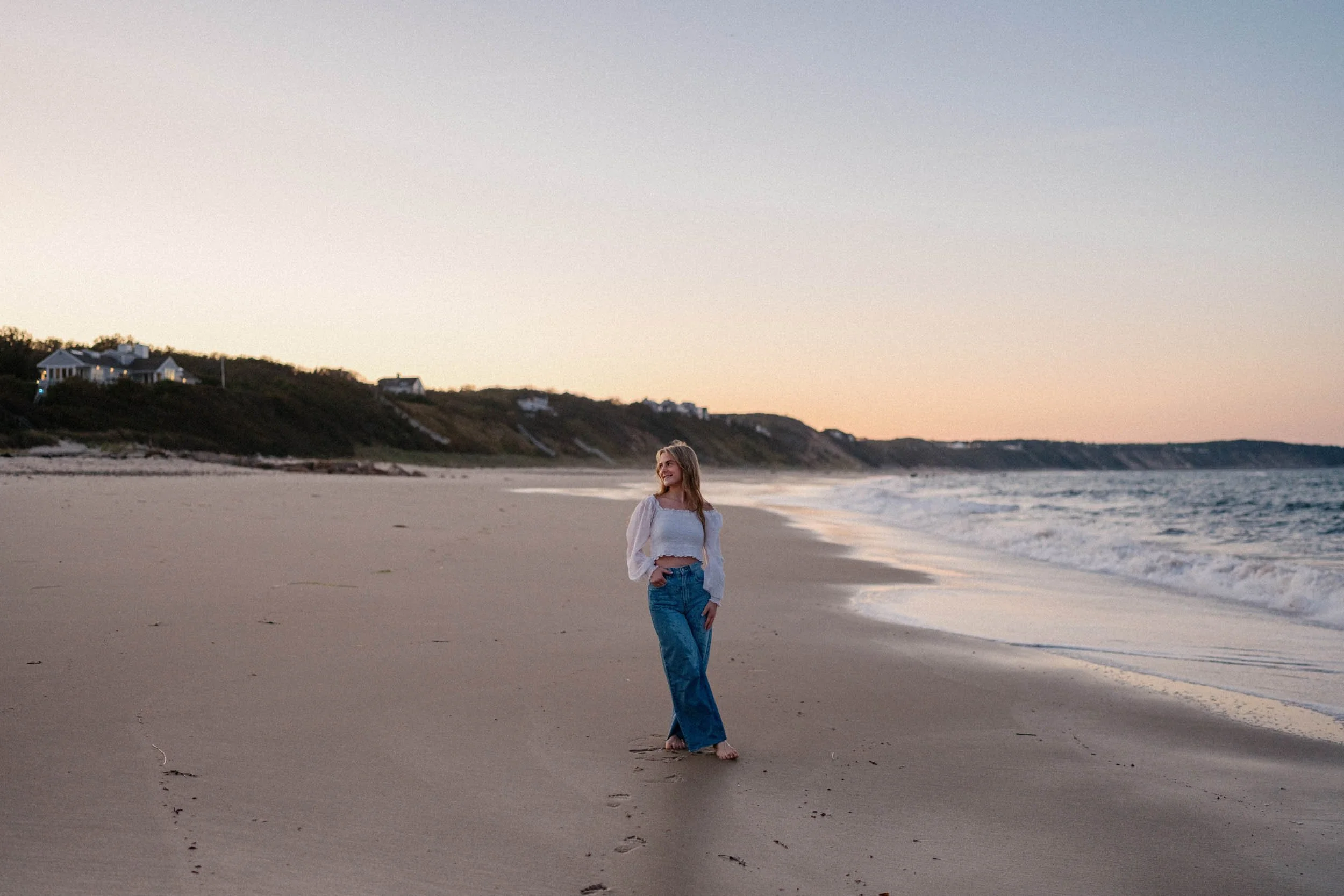 A woman standing on a sandy beach at sunset, facing the ocean with a smile, wearing a white crop top, blue jeans, and a light-colored shirt, with houses on a hill in the background.