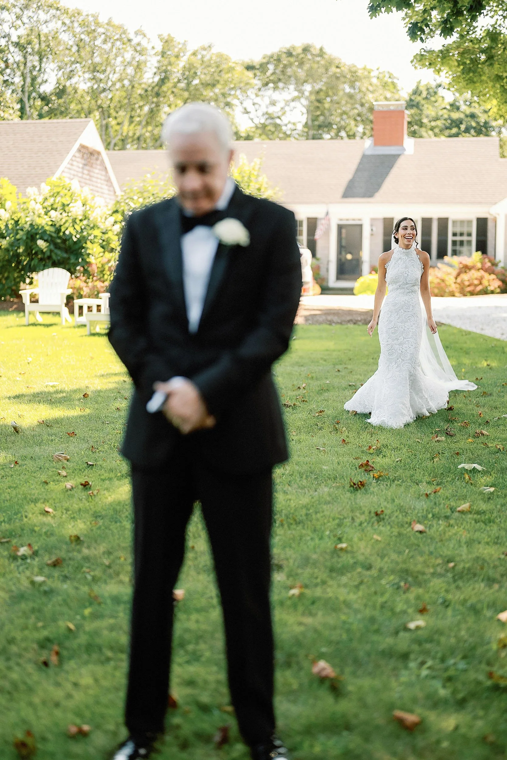 A bride in a white lace wedding gown smiling in the background, while an older man in a black tuxedo with a white boutonniere stands in the foreground with his head bowed.