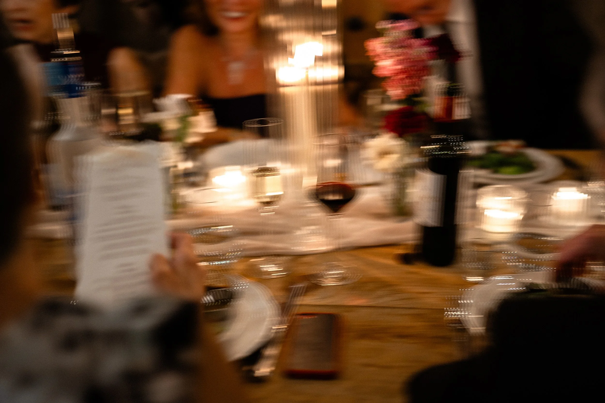 Blurred image of a dinner party with people sitting around a table with candles, wine glasses, and floral centerpieces
