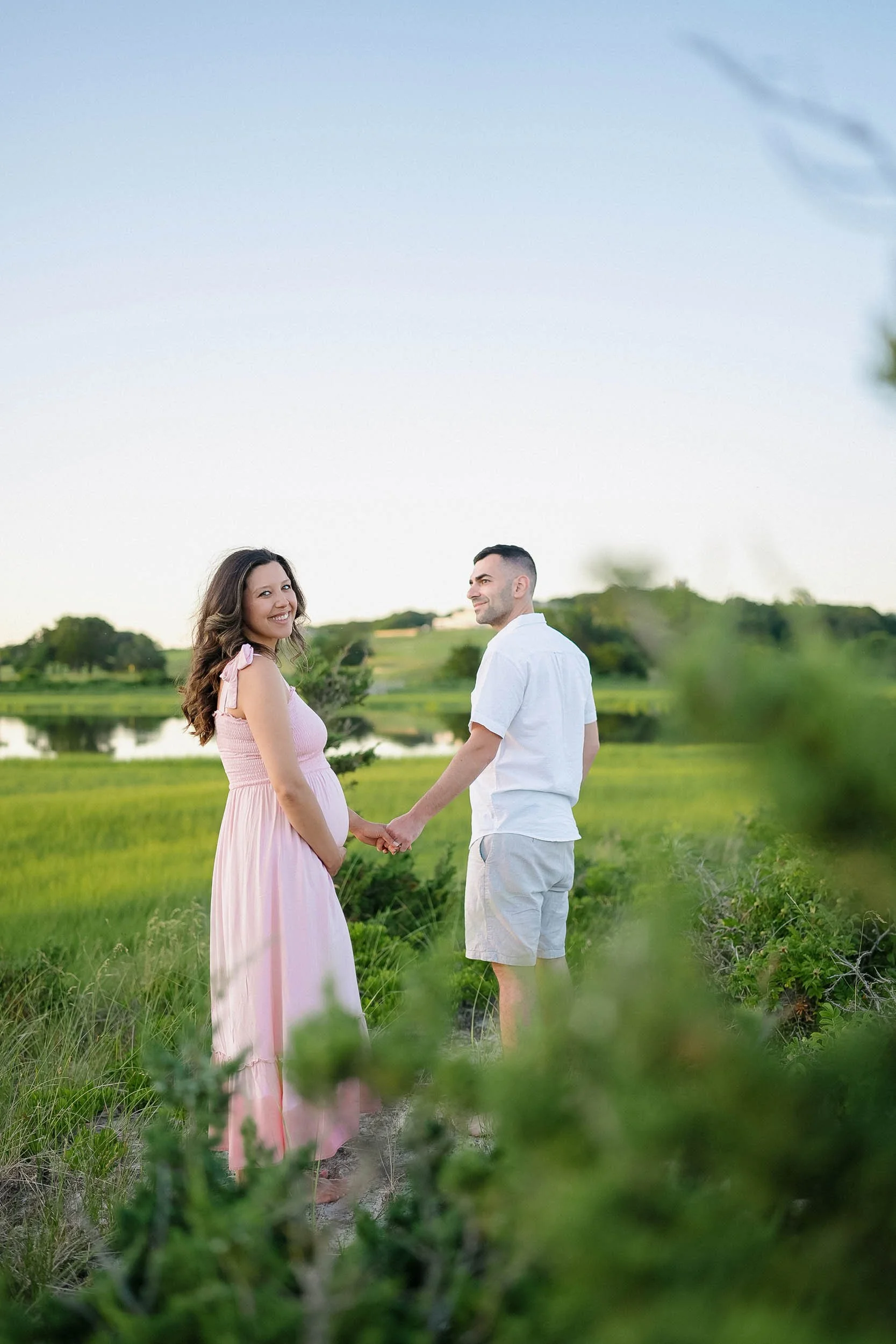 A pregnant woman in a pink dress holding hands with a man in a white shirt and shorts, standing outdoors on a grassy field with a lake and trees in the background. They are smiling and looking at each other.