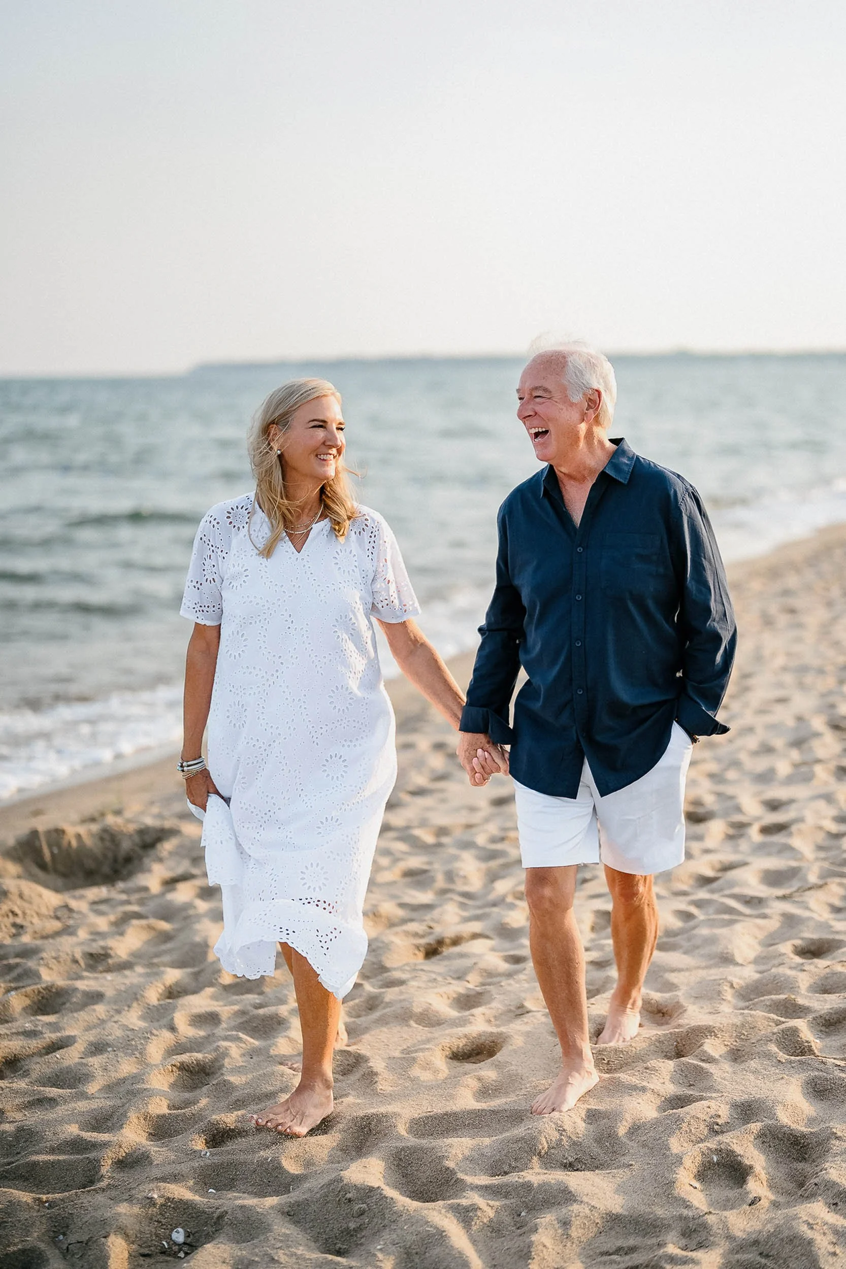An elderly couple walking on the beach holding hands, smiling, and enjoying each other's company.