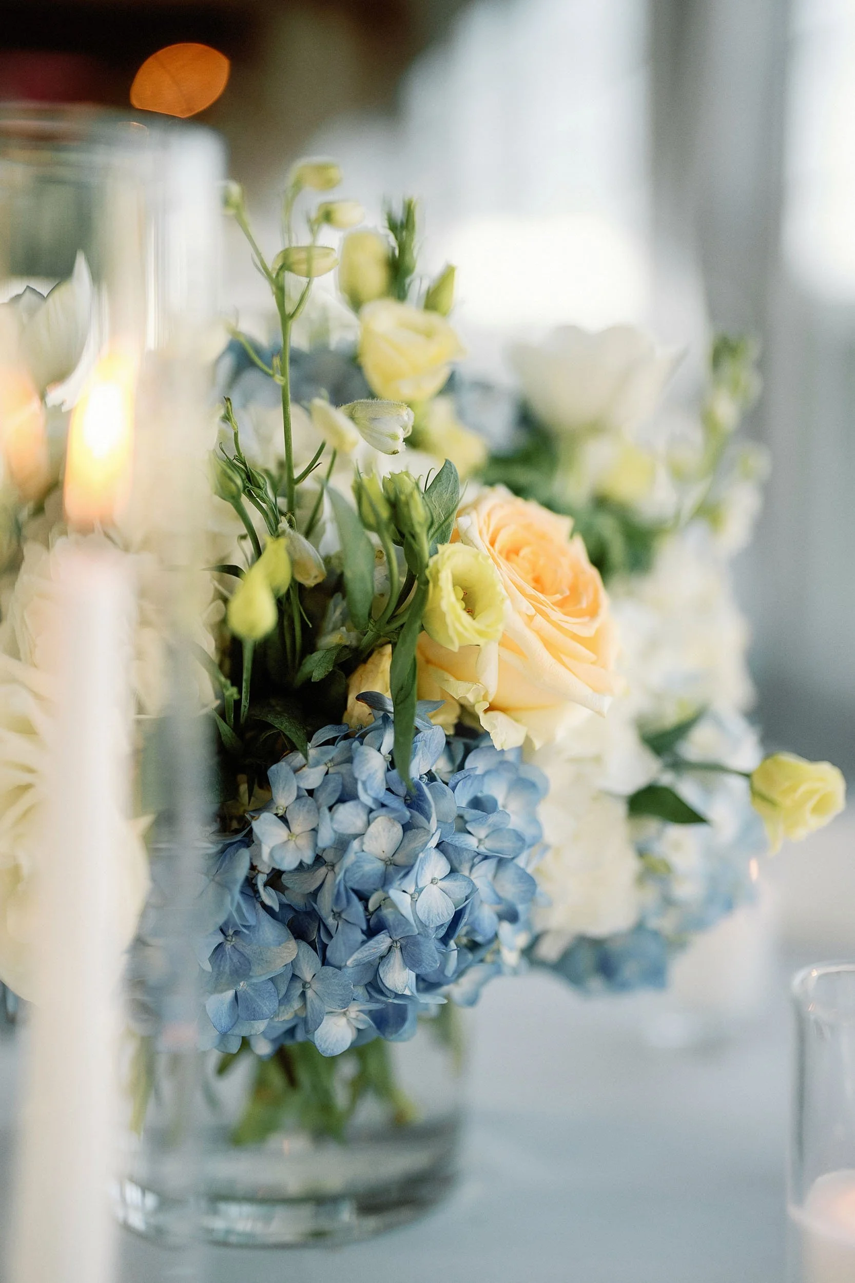 A close-up of a floral arrangement with light yellow roses, blue hydrangeas, and white flowers in a glass vase.