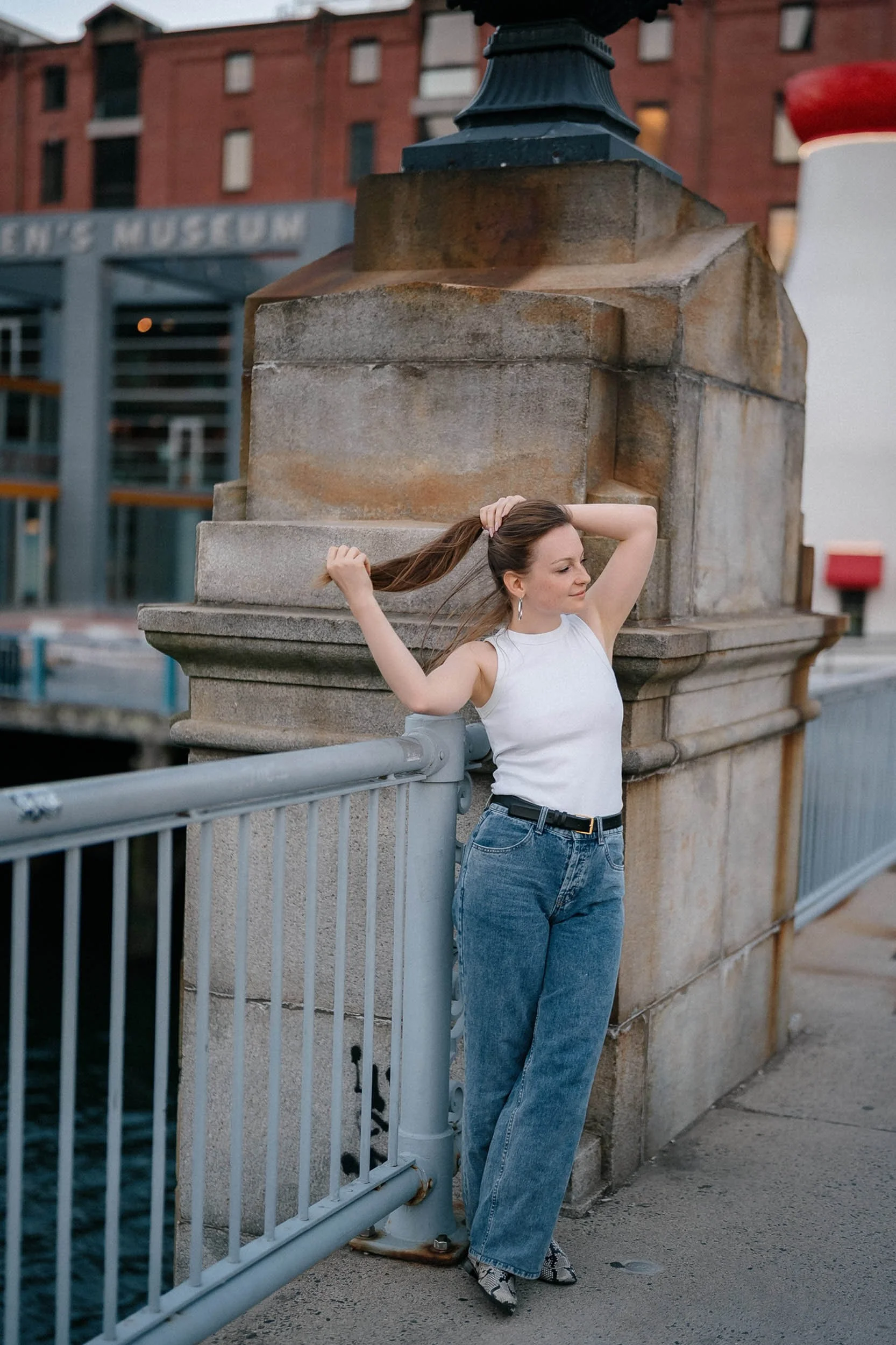 A woman standing on a bridge, adjusting her hair, with an urban backdrop that includes a building with a sign reading 'Museum'.