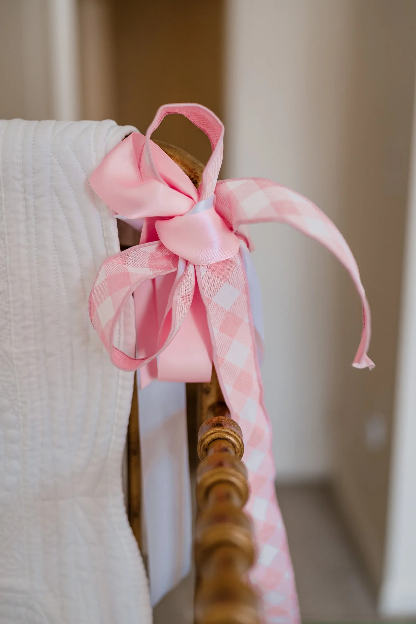 Close-up of a pink and checkered ribbon tied in a bow on the corner of a wooden bed or crib, with part of a white quilted fabric visible.