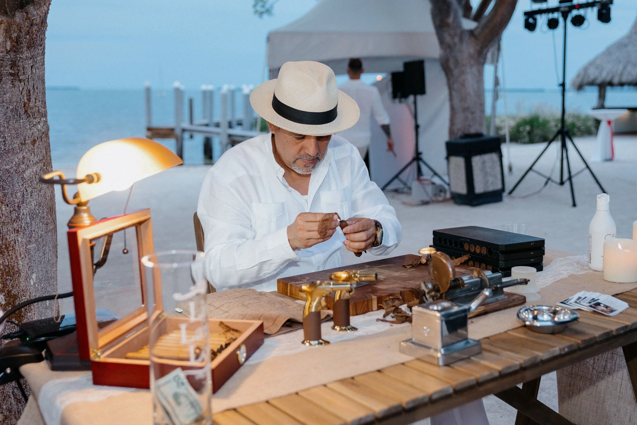 A man wearing a white shirt and a straw hat sits at a wooden table outdoors by the water, working with cigar tools. The scene includes a lamp, a glass of water, and various cigar accessories with a backdrop of trees, water, and a tent or canopy.