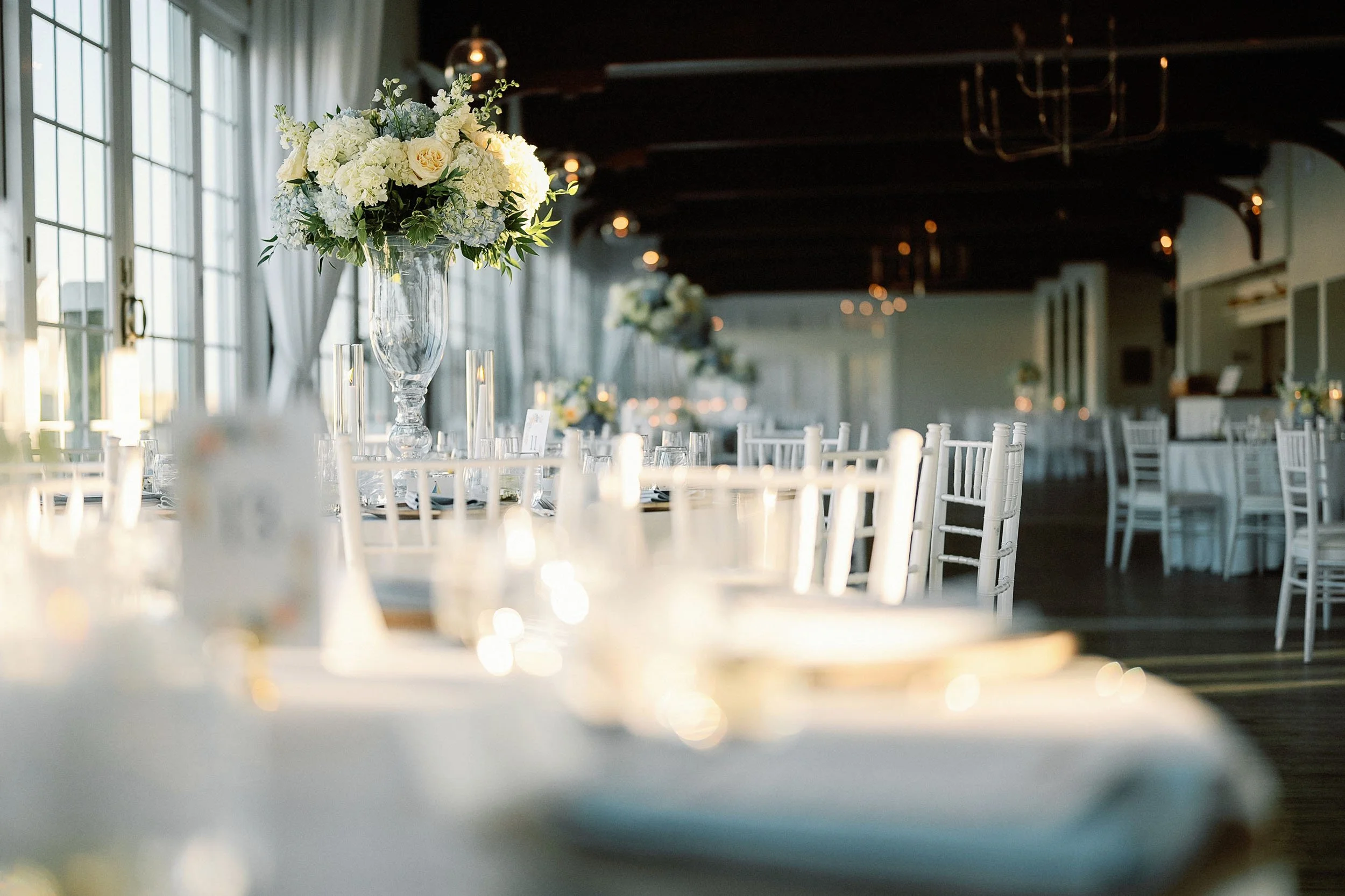 Elegant banquet hall decorated for a wedding or special event with white chairs, a tall floral centerpiece with white and light yellow flowers in a clear glass vase, and soft natural light coming through large windows.