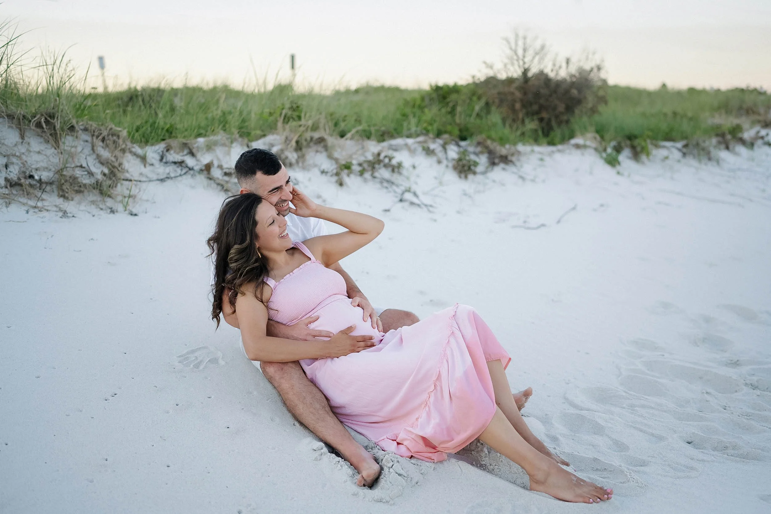 A couple sitting on the sandy beach, with the woman pregnant, smiling and enjoying each other's company.