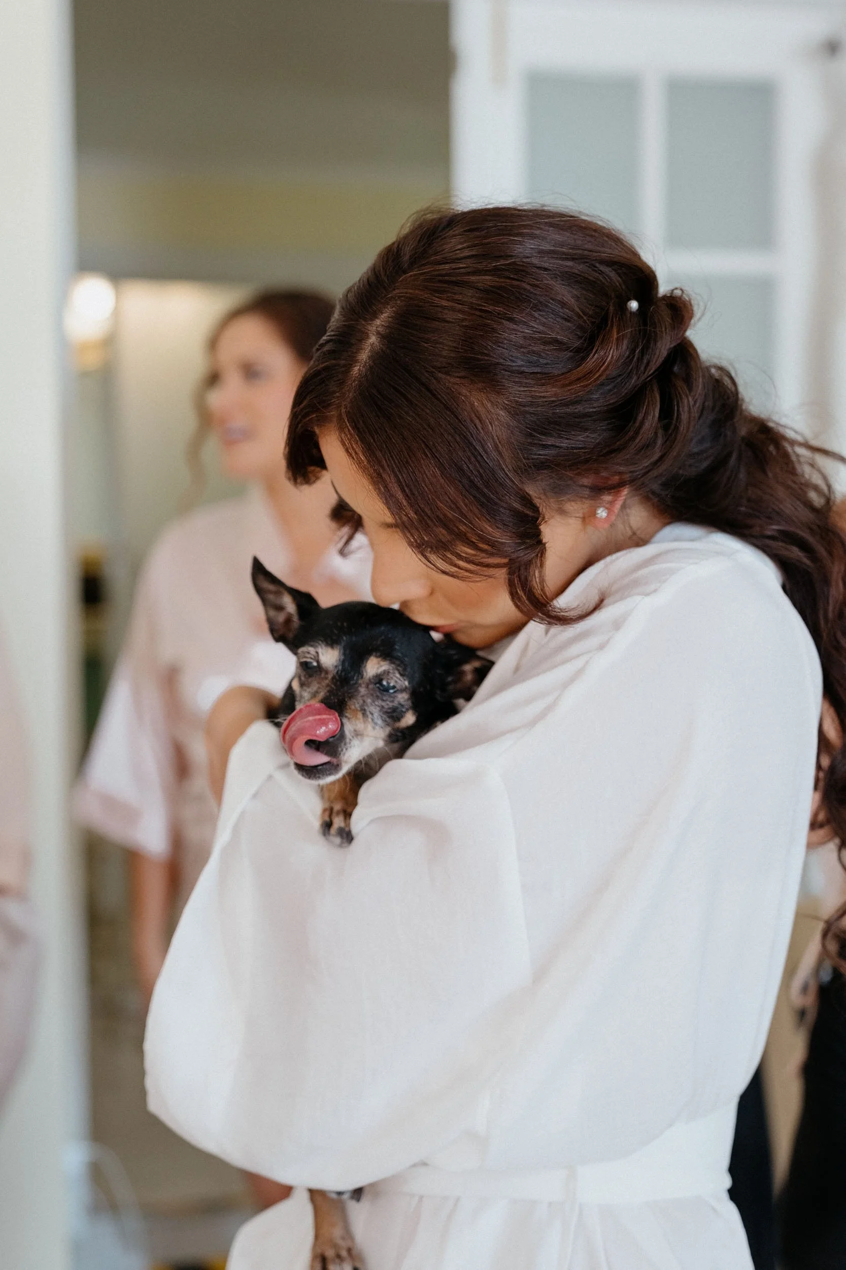 A woman in a white garment holding a small black and tan dog, which is licking its nose, in a bright indoor setting.