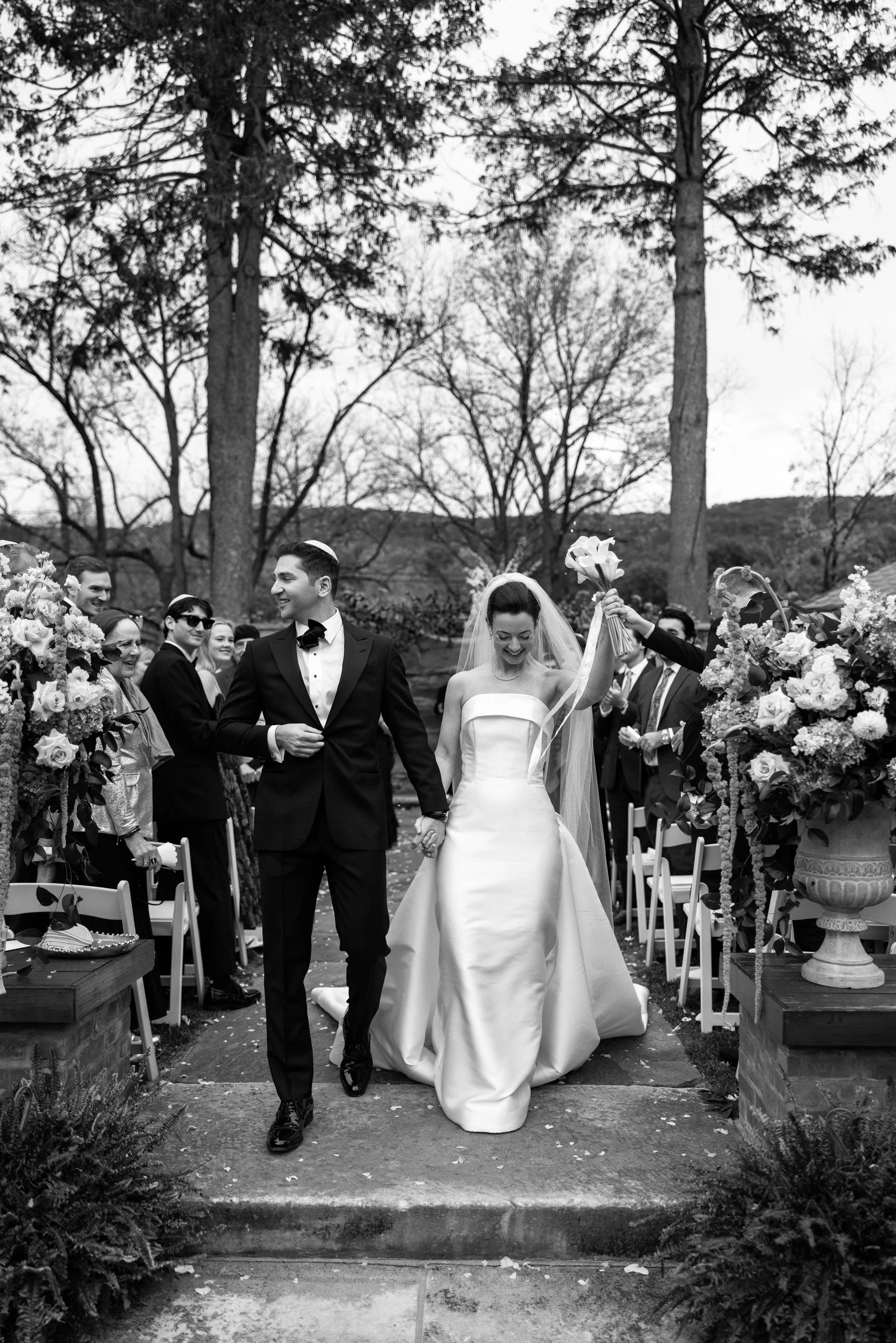 A black-and-white photo of a bride and groom walking down the aisle after their wedding ceremony outside, surrounded by guests and large flower arrangements.