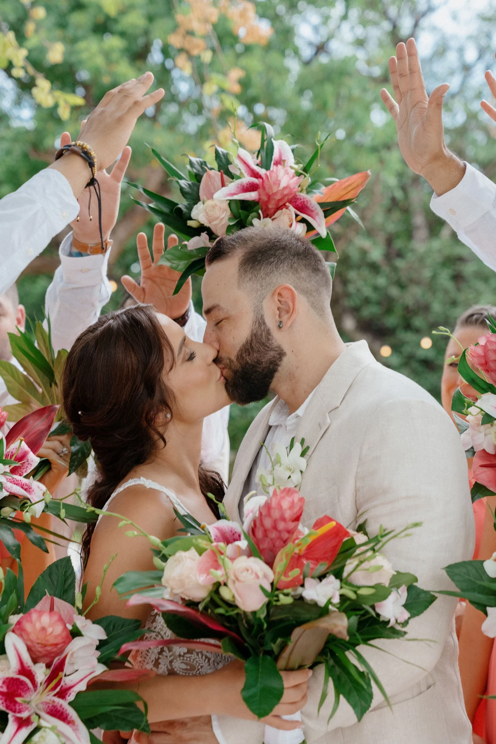 A bride and groom kiss during their wedding ceremony, surrounded by friends holding up flowers and greenery.