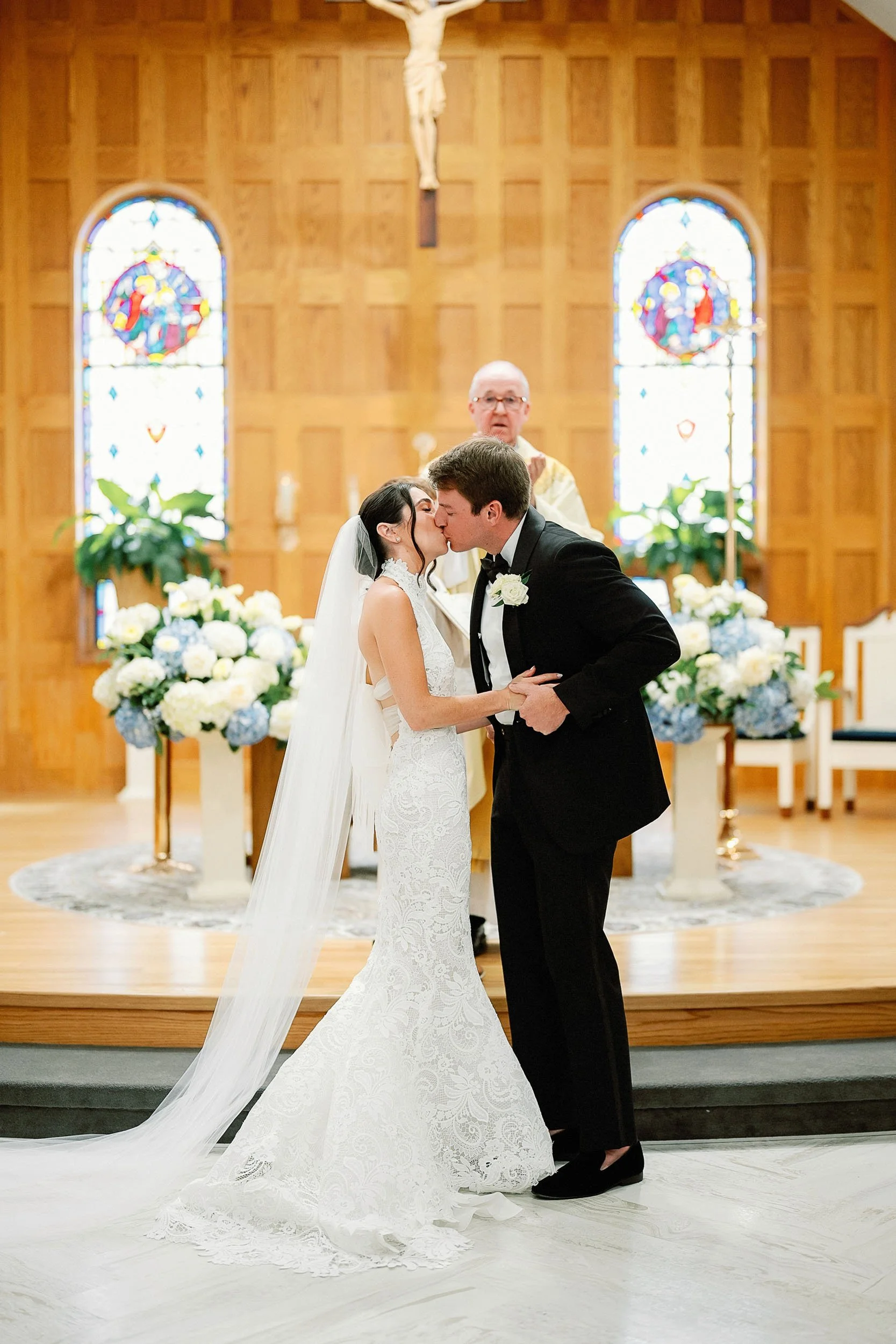 A bride and groom kiss at their wedding ceremony in a church, with a priest in the background, stained glass windows, and floral arrangements.