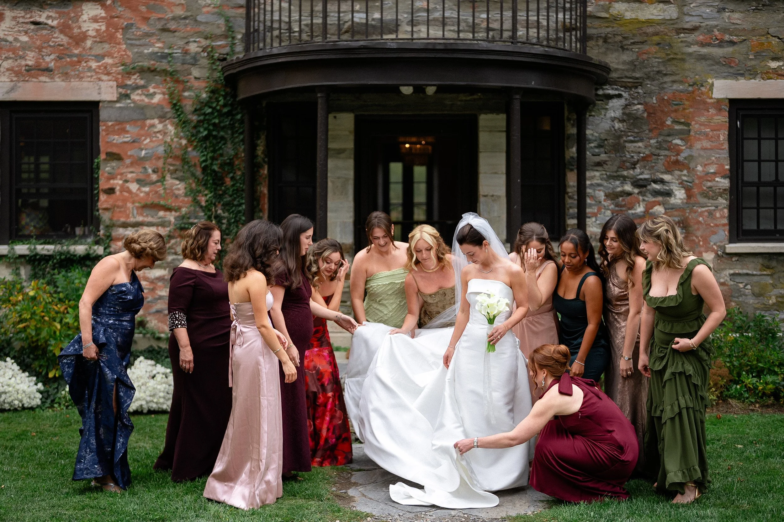 A bride in a white wedding gown holding a bouquet of white calla lilies is surrounded by women in formal dresses, all looking down at her dress in front of a stone building with black windows and a decorated garden.
