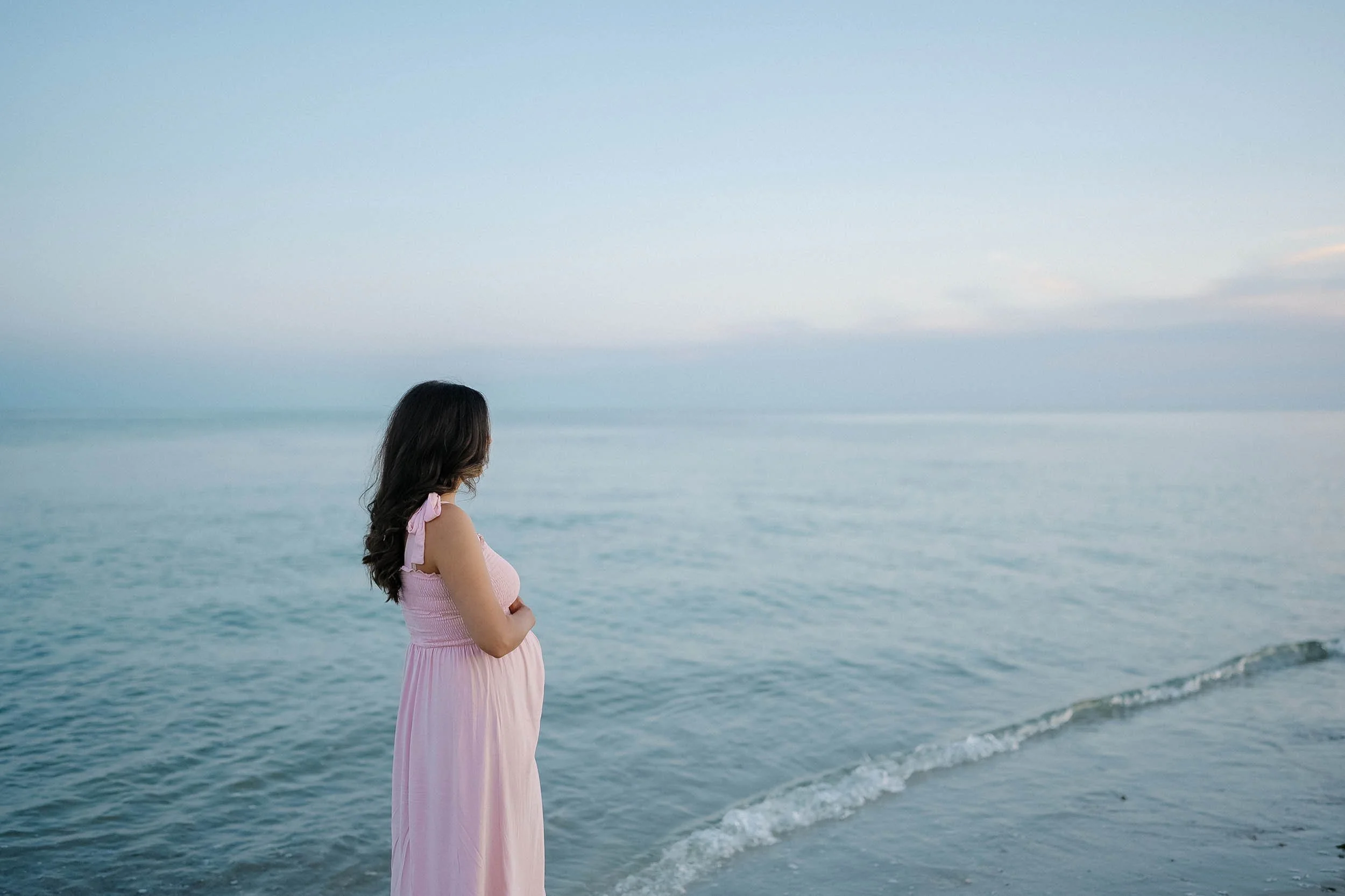A woman in a pink dress stands on a beach looking at the ocean during sunset.