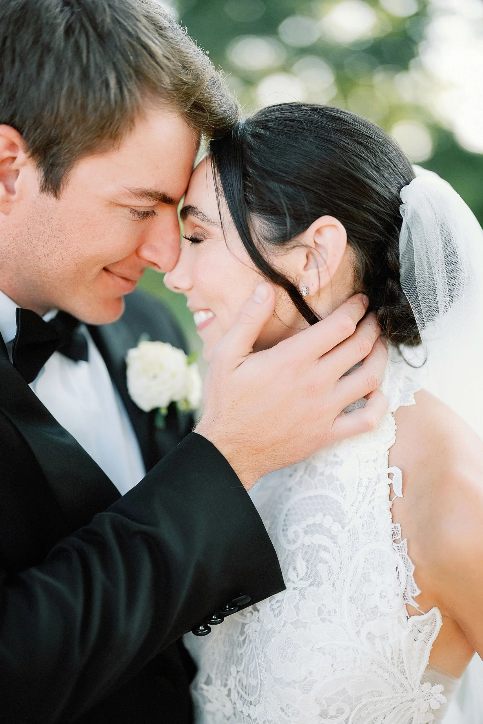 A newlywed couple with their foreheads touching and eyes closed, smiling, outdoors in wedding attire, with a blurred green background.