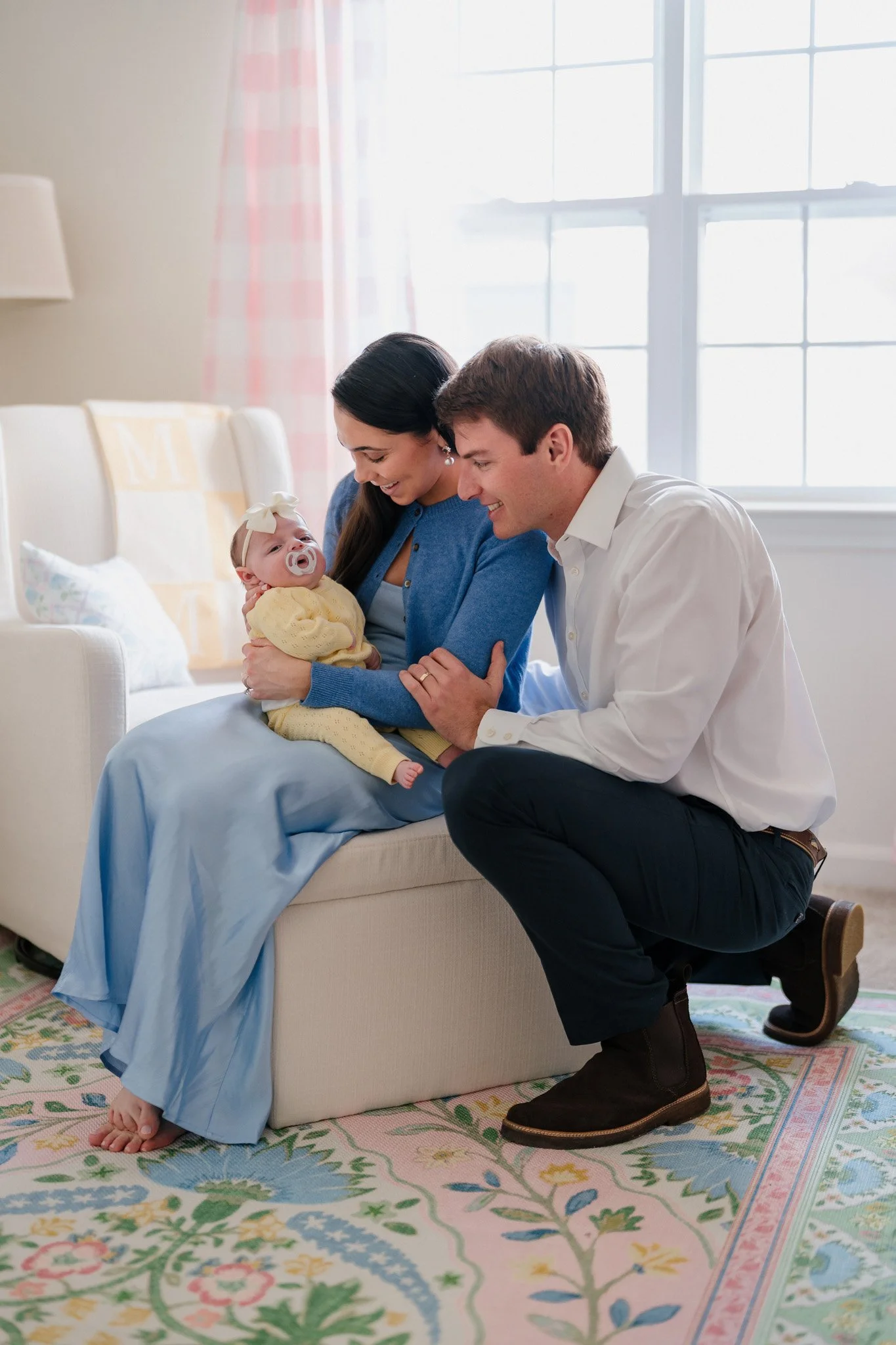 Family with baby sitting on couch, mother holding baby with pacifier, father kneeling beside them, all smiling in a bright living room.