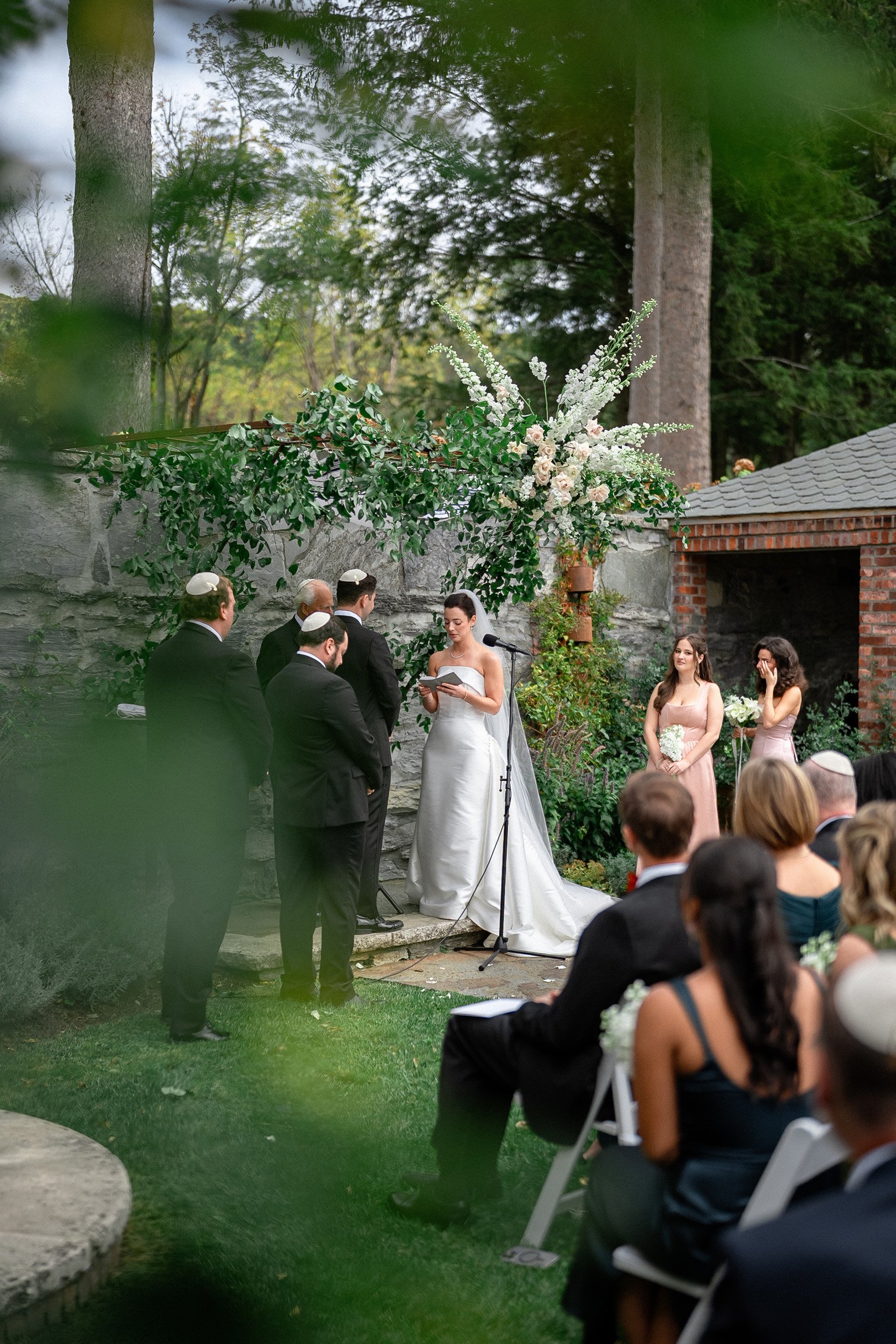 A wedding ceremony outdoors with a bride and groom reading vows, officiant, bridesmaids, groomsmen, and guests seated in chairs, decorated with large floral arrangements and surrounded by trees.