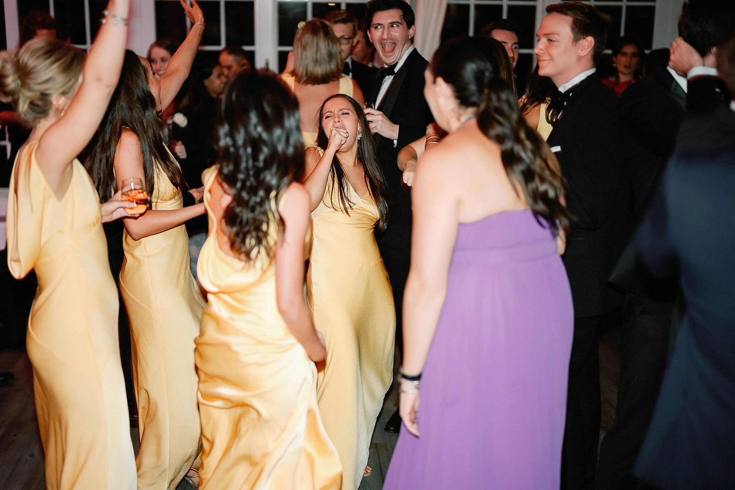People dancing and enjoying themselves at a formal event or wedding reception, some women in yellow dresses, men in black tuxedos.