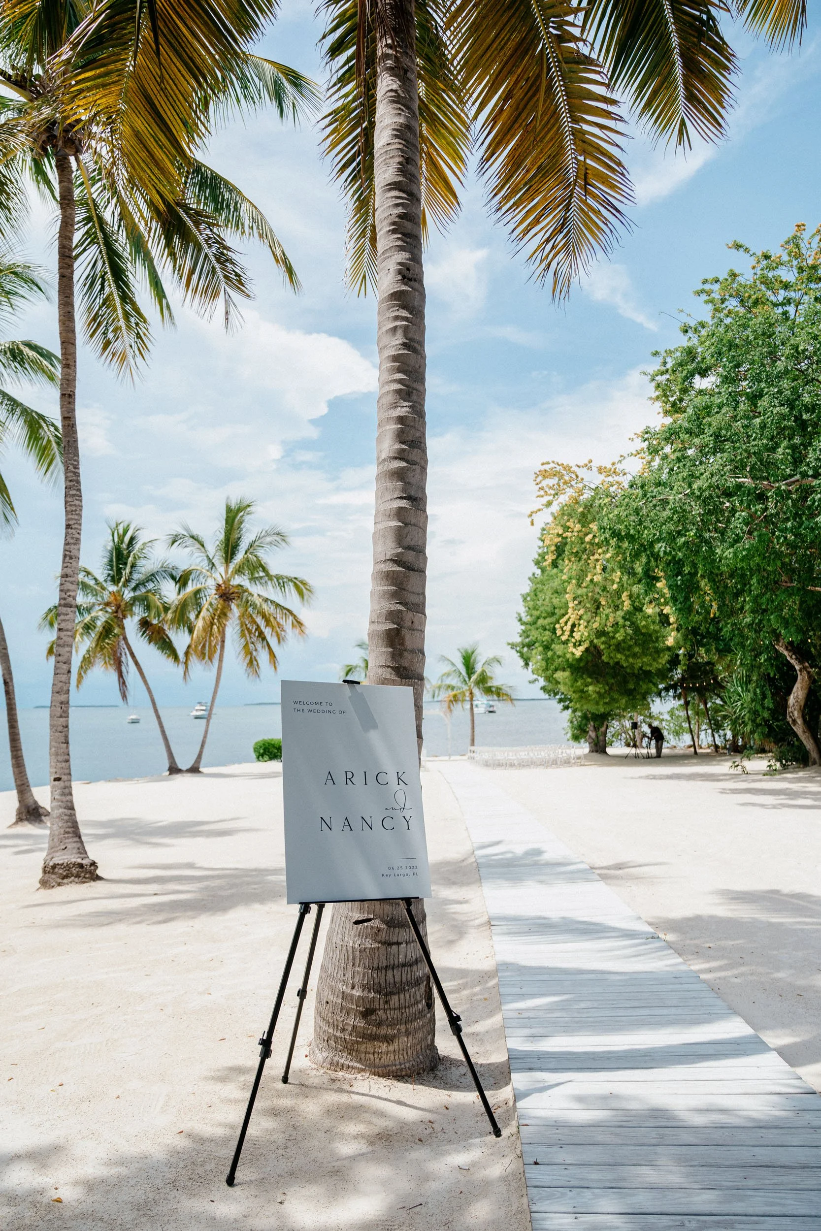 A wedding welcome sign on a beach pathway, with palm trees and the ocean in the background, indicating a wedding at Key Largo, Florida.