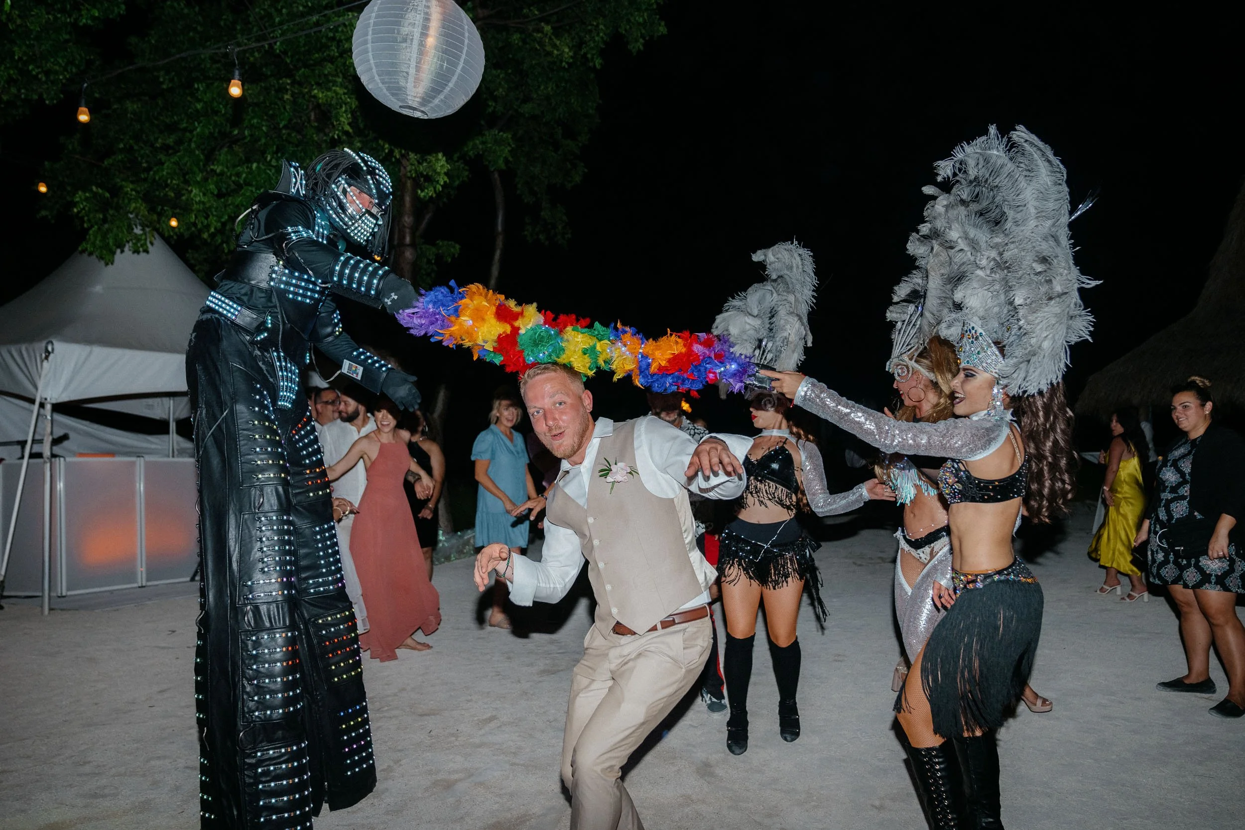 People dancing at a nighttime outdoor event, with two performers in elaborate costumes and feathered headpieces, holding a colorful feather boa.