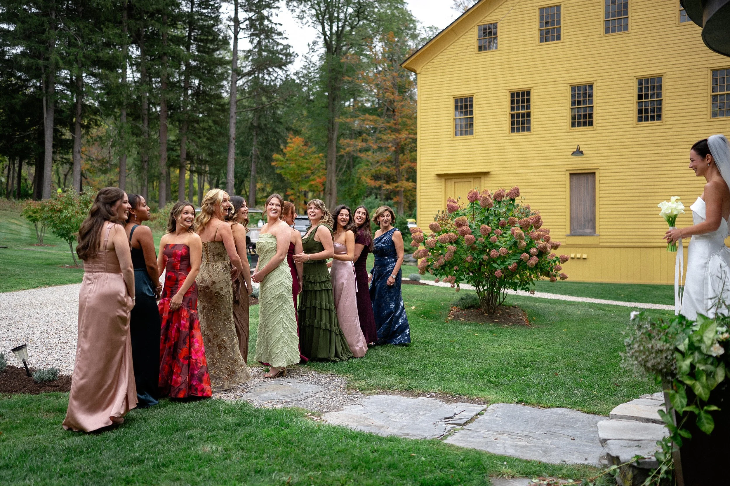 A bride in a white dress holding a bouquet stands in front of a group of women in colorful dresses at a wedding outdoor ceremony, with a yellow building and trees in the background.