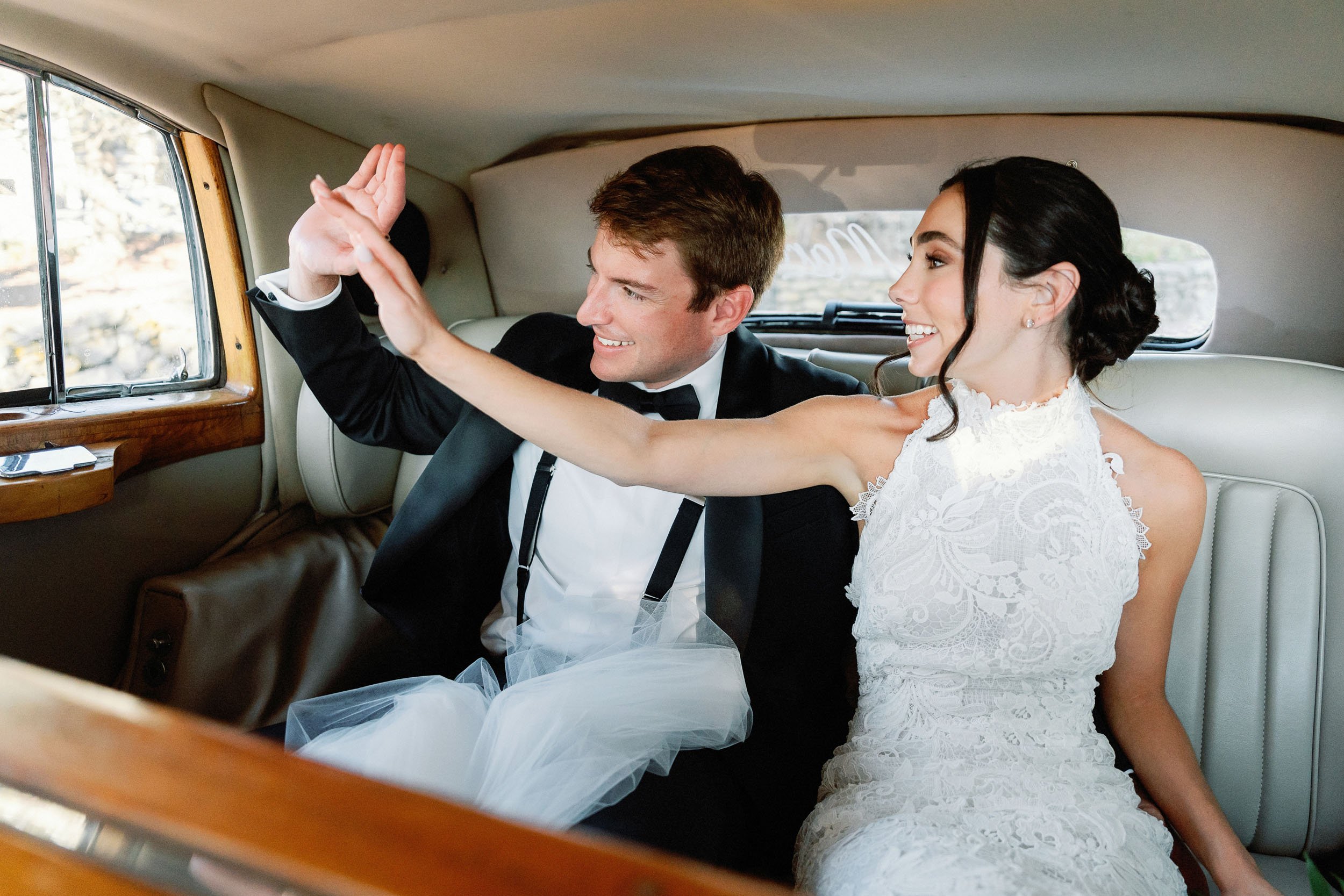 Happy bride and groom sitting inside a vintage car, sharing a high-five or celebratory gesture after their wedding.