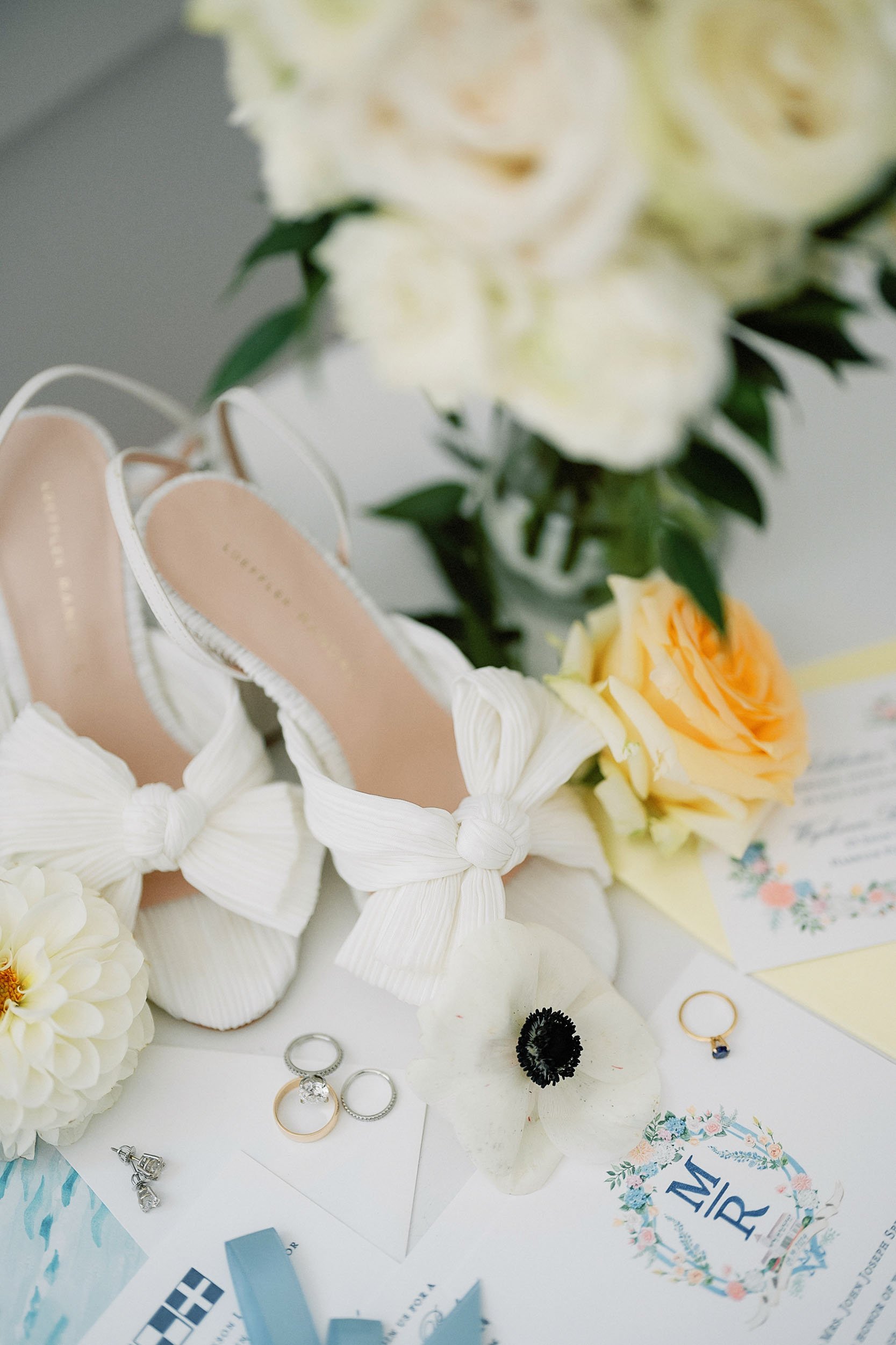 Wedding flat shoes with bows, surrounded by flowers, rings, and wedding invitations.