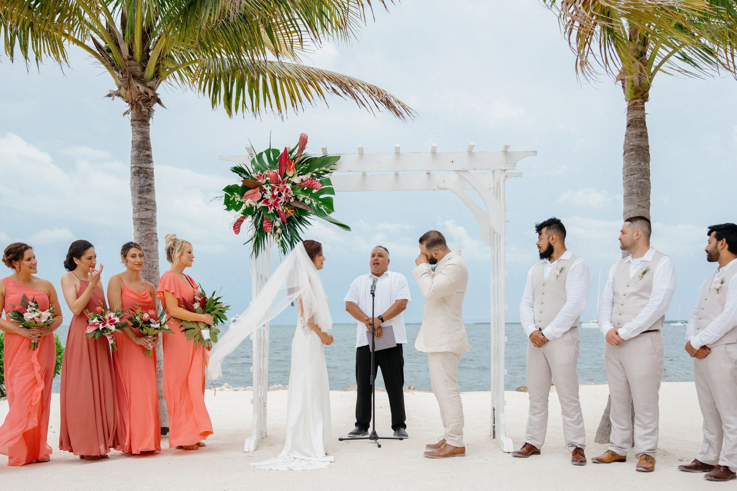 Beach wedding ceremony with bride and groom, officiant, bridesmaids, and groomsmen under a white wooden arch decorated with pink and green tropical flowers, with two palm trees and the ocean in the background.