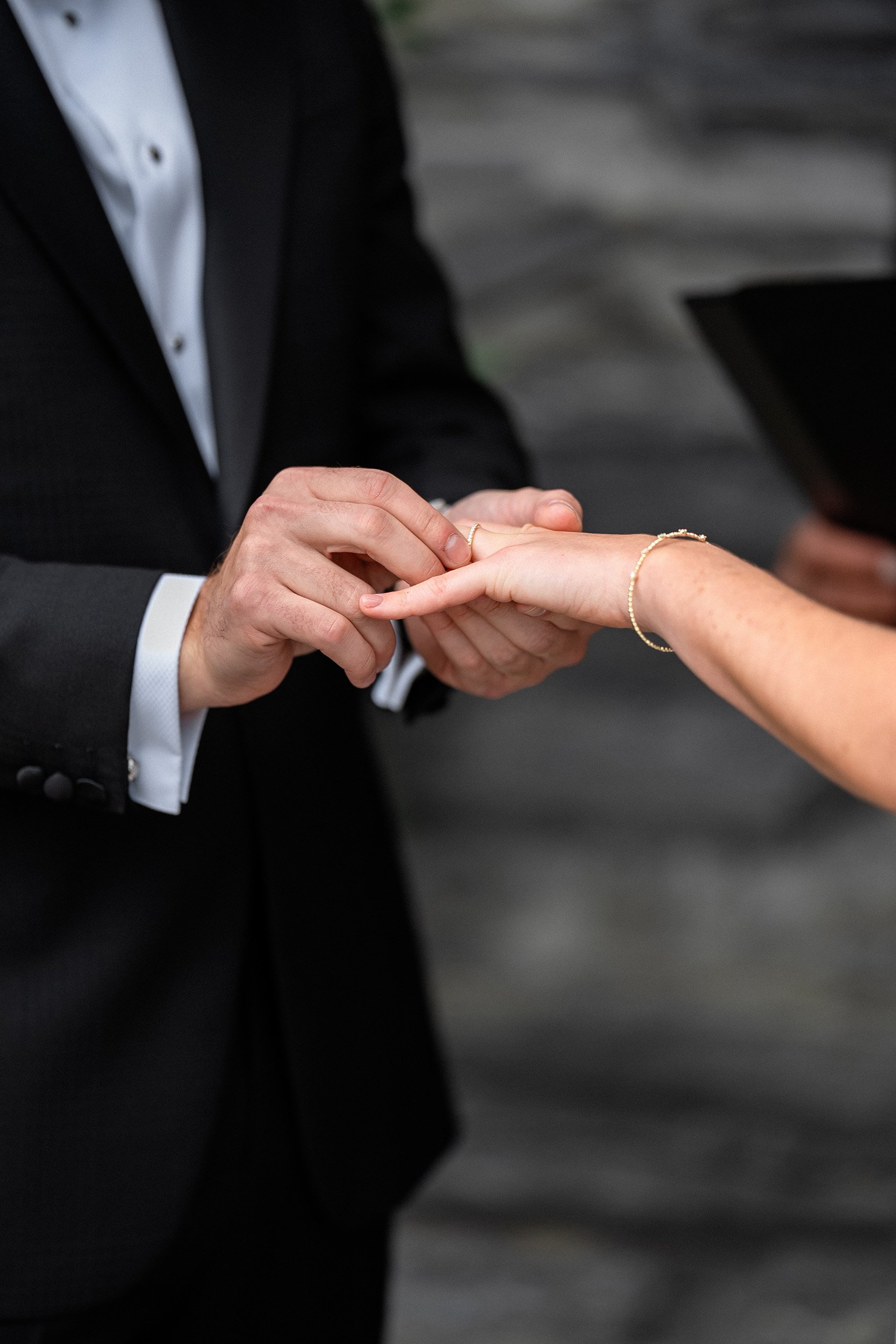 Person in a black suit and white shirt placing a wedding ring on another person's finger.