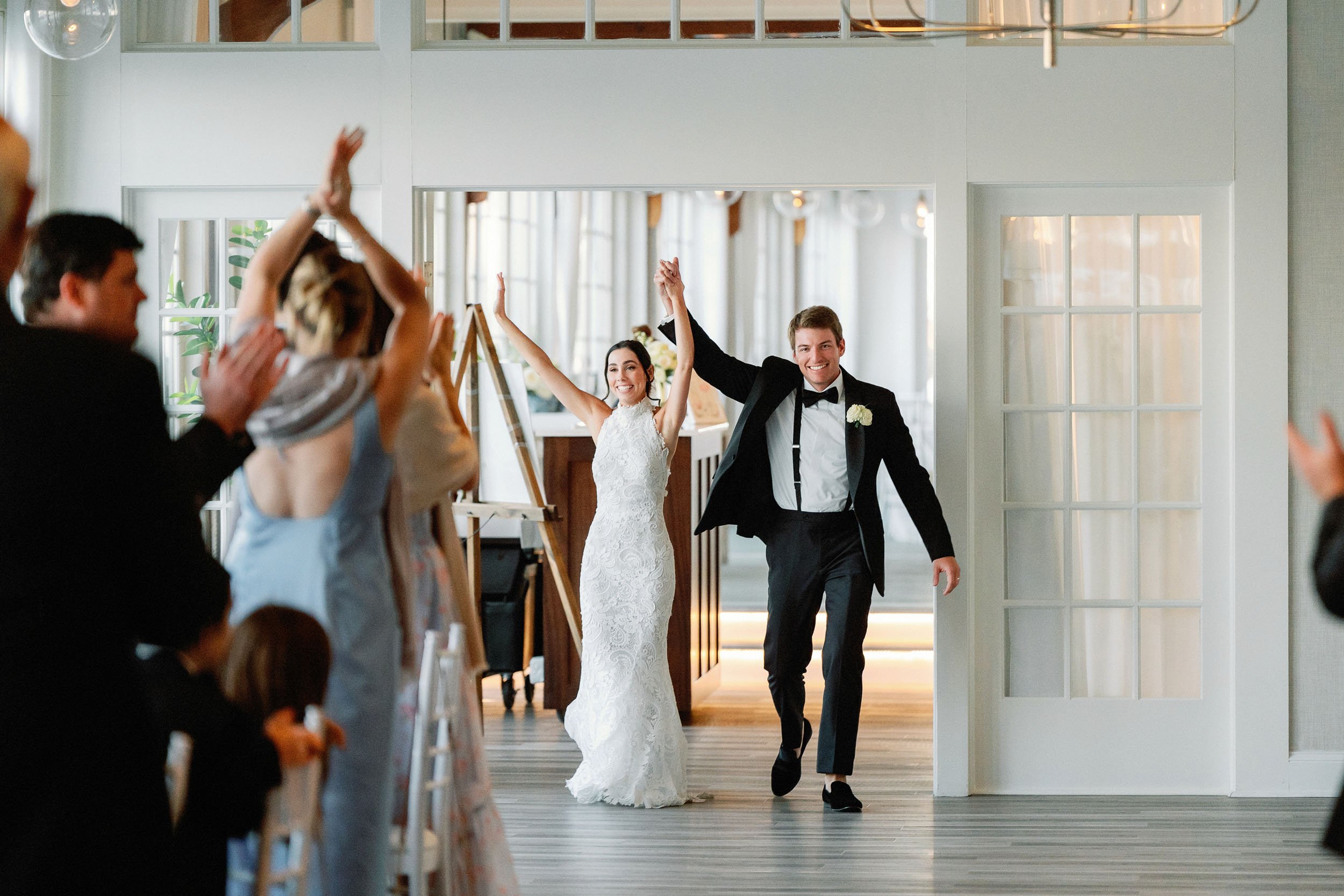 Bride and groom walking into wedding reception, smiling and raising hands in celebration, with guests clapping and cheering.