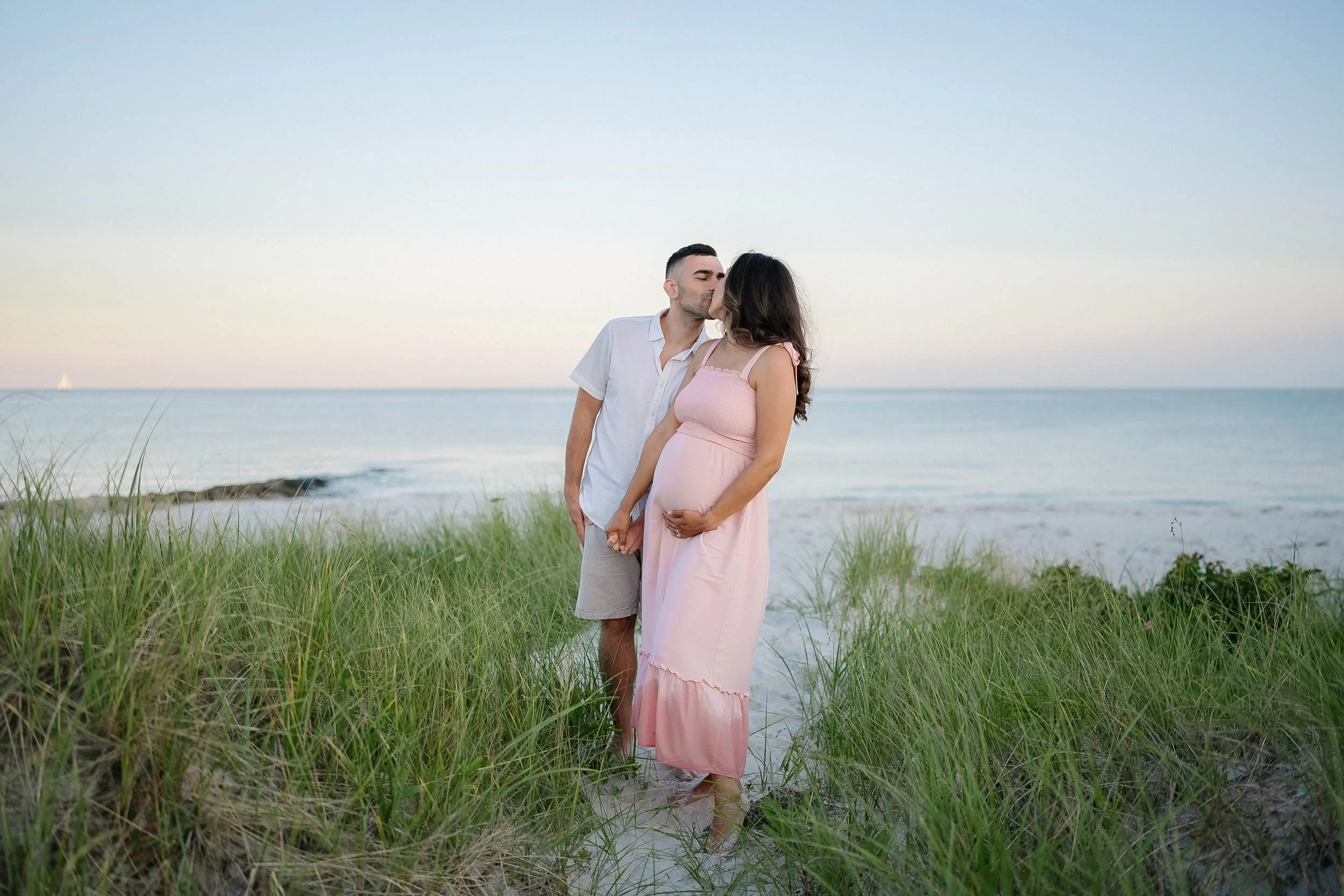 A man and a pregnant woman kissing on a beach with grass and ocean in the background.