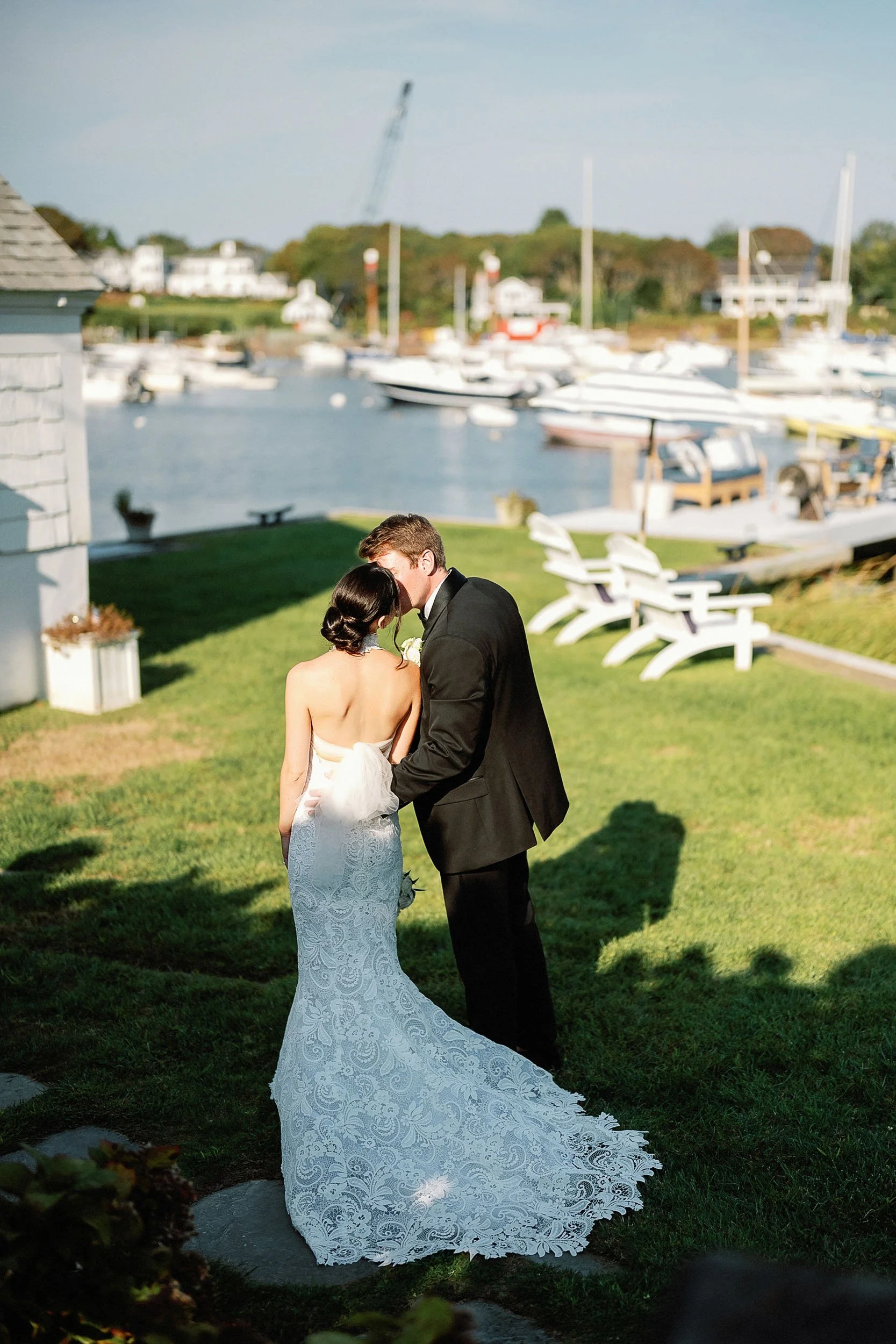 A bride and groom share a romantic kiss outdoors near a marina, with sailboats docked in the background.