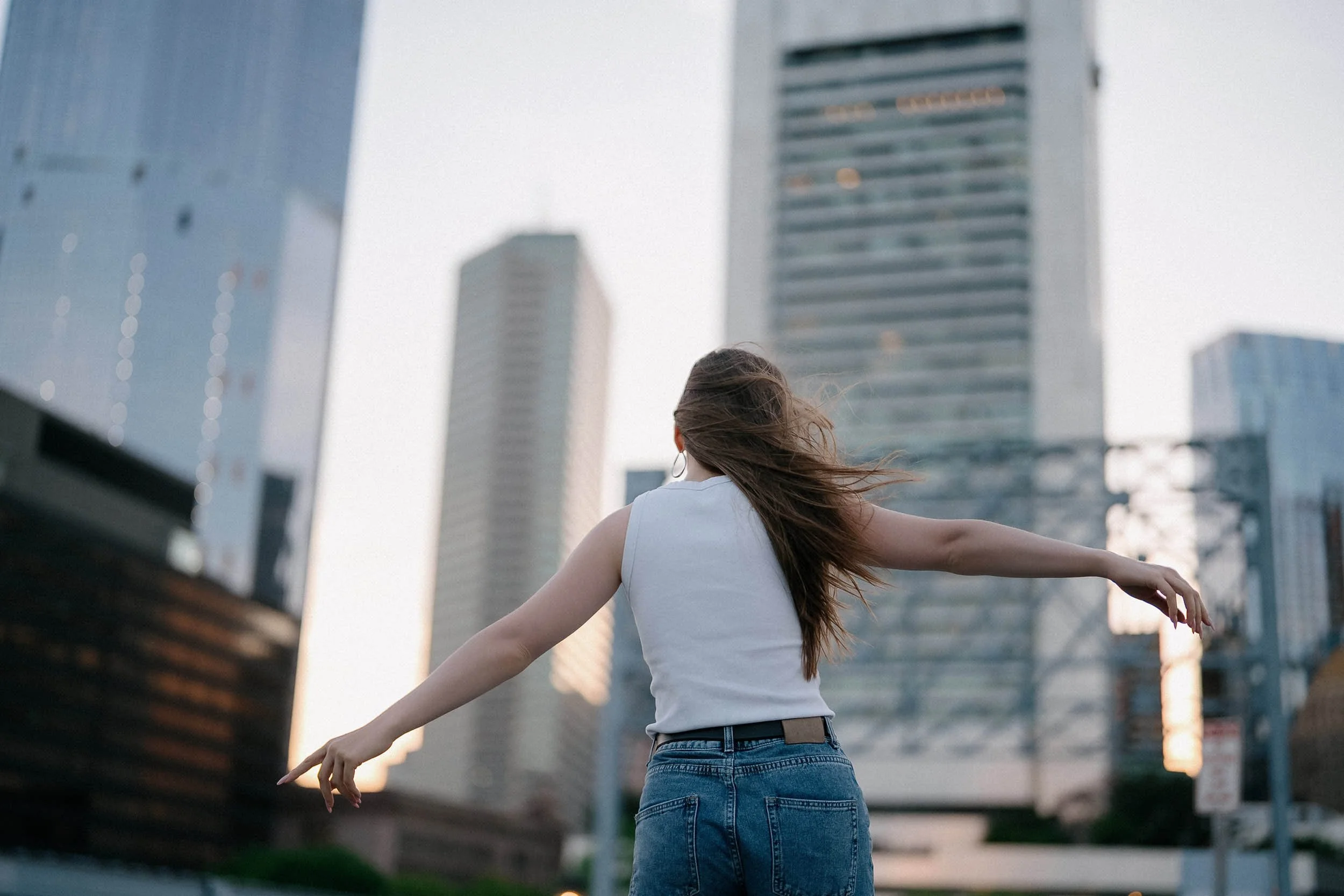 A woman with long hair wearing a white sleeveless top and blue jeans standing outdoors with arms outstretched against a backdrop of tall city buildings.