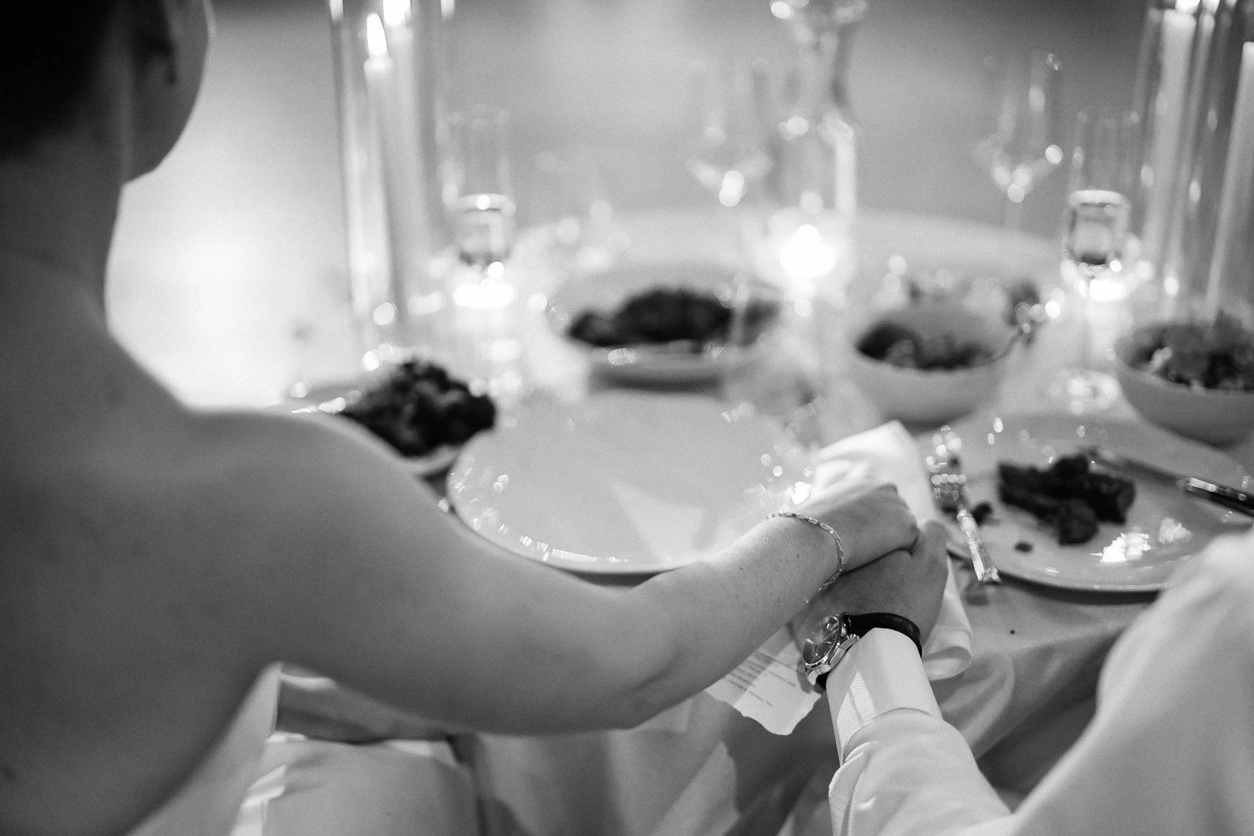 A couple sitting at a dining table holding hands during a meal, with desserts, glasses, and plates on the table.