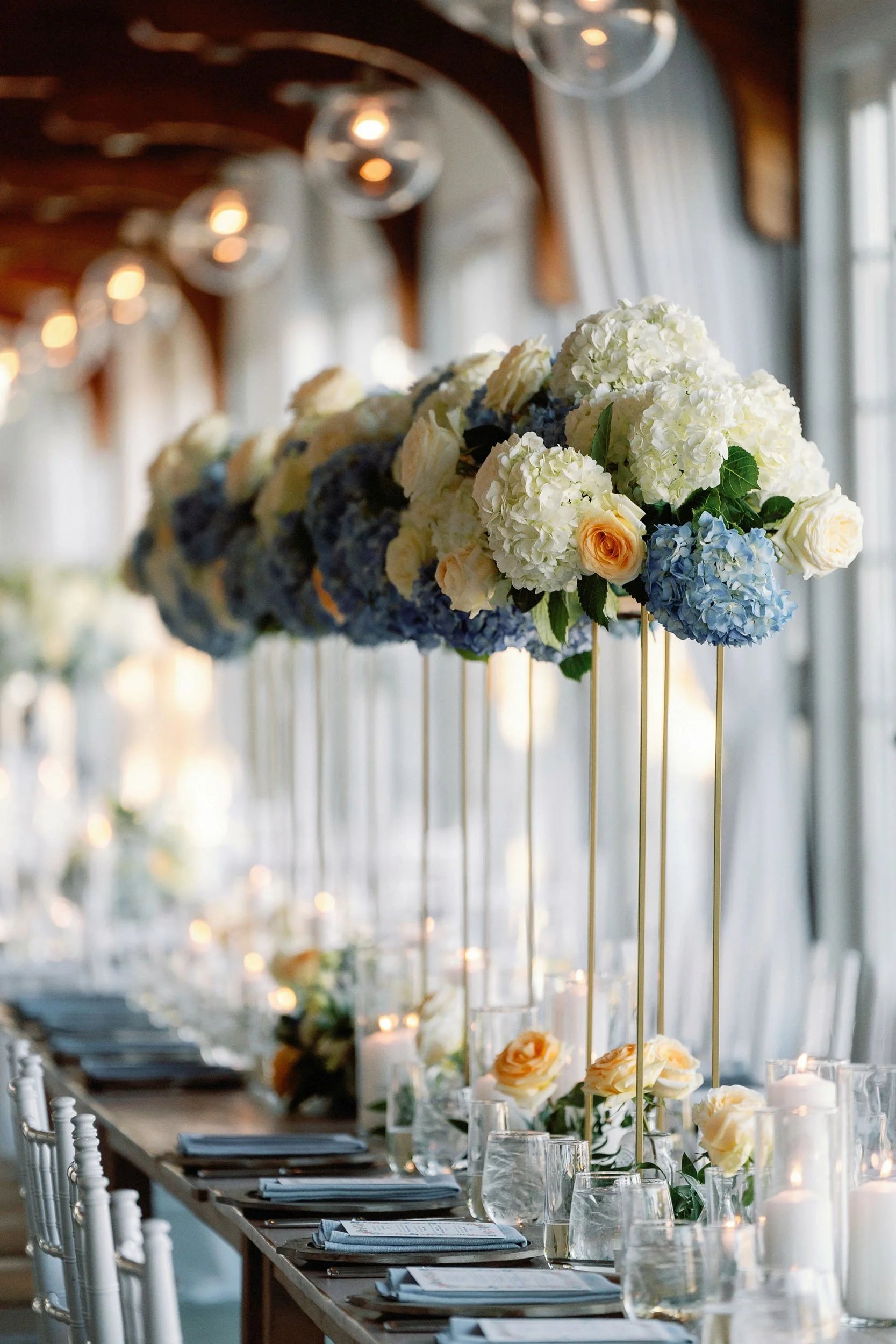 Wedding table with large floral centerpiece featuring white, blue, and yellow roses, hydrangeas, and greenery, set with candles, glasses, and silverware.
