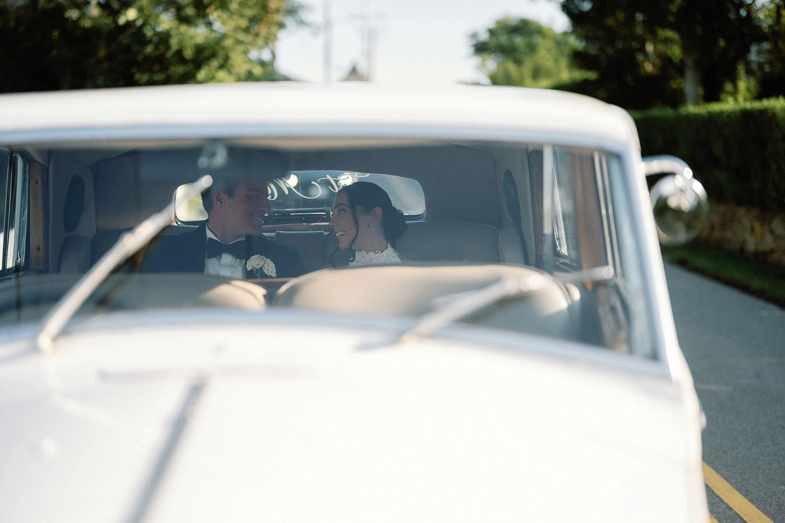 A bride and groom sitting inside a vintage white car, smiling and looking at each other, on a street with greenery and trees in the background.