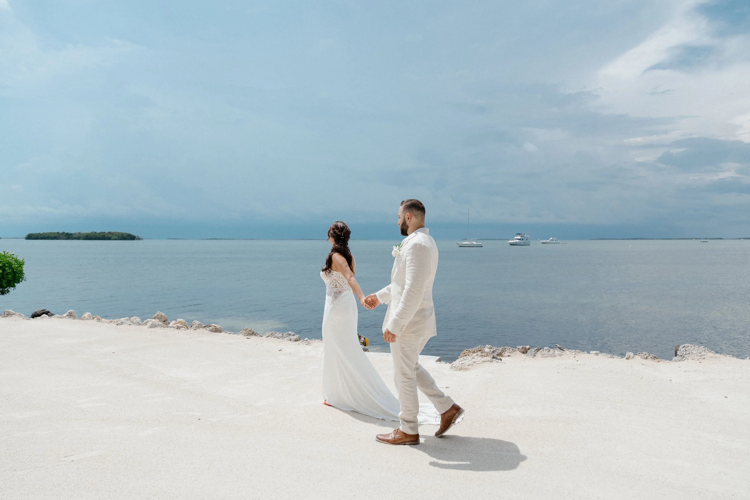 A man and woman dressed in wedding attire holding hands on a beach with the ocean and boats in the background.