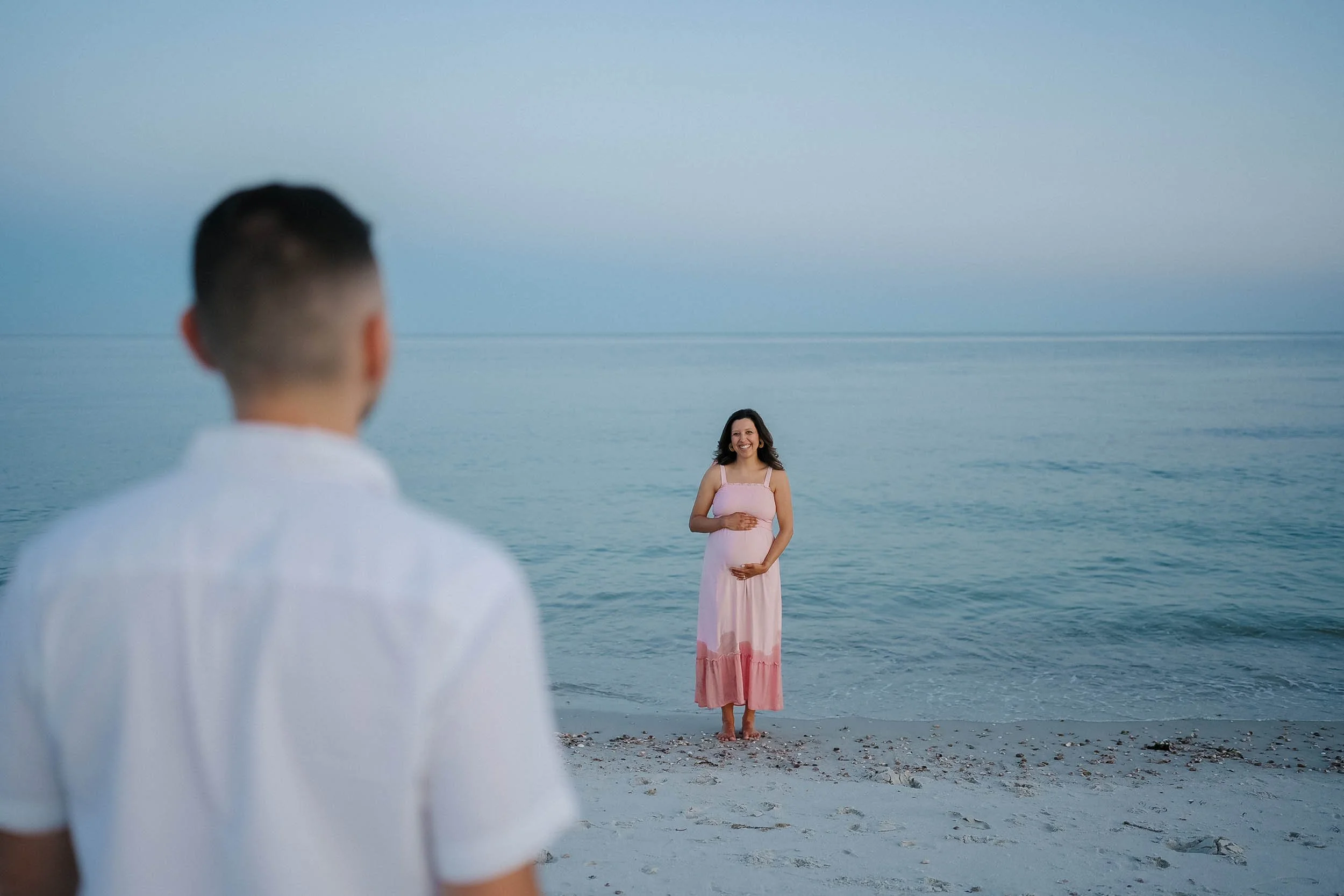 A woman in a pink dress standing on a beach, about to have a photoshoot with a man in a white shirt, with the ocean behind them.